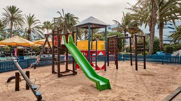 Kids can play on the green slide surrounded by palm trees and a sandy ground.