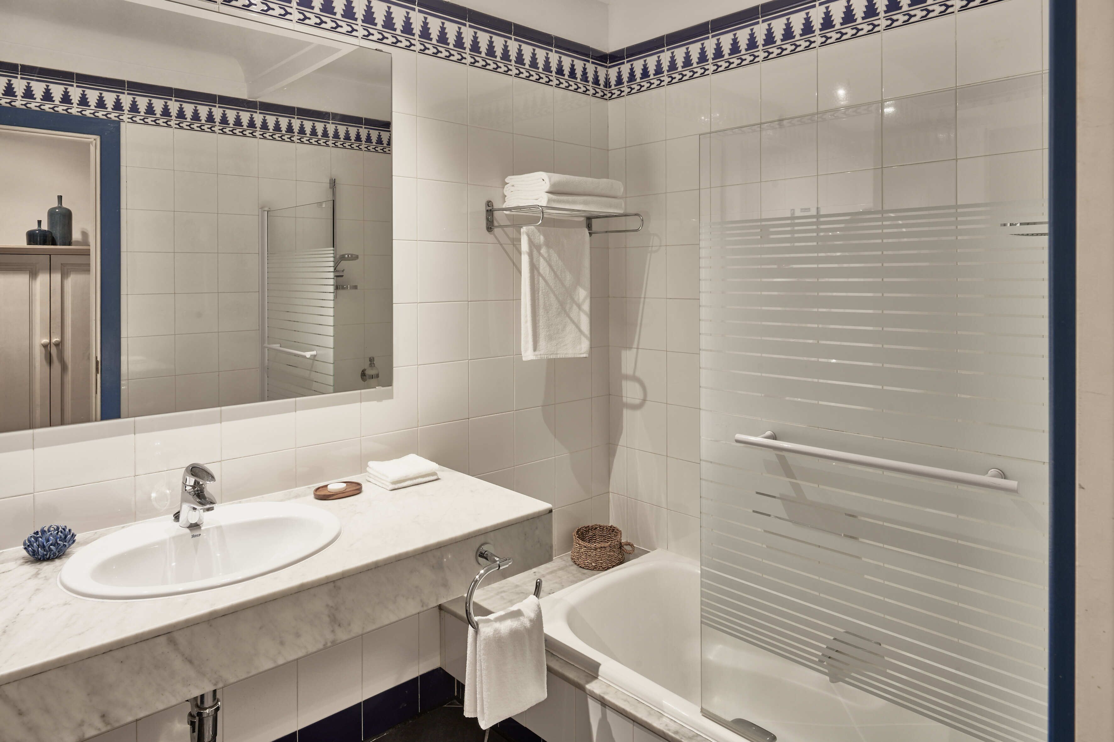 A bathroom with a white sink, mirror, and bathtub. There is a towel rack with towels and a basket.