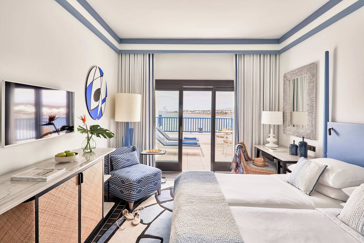Hotelkamer met een bed, een bureau met een televisie, een balkon met zwembad en uitzicht op het strand.