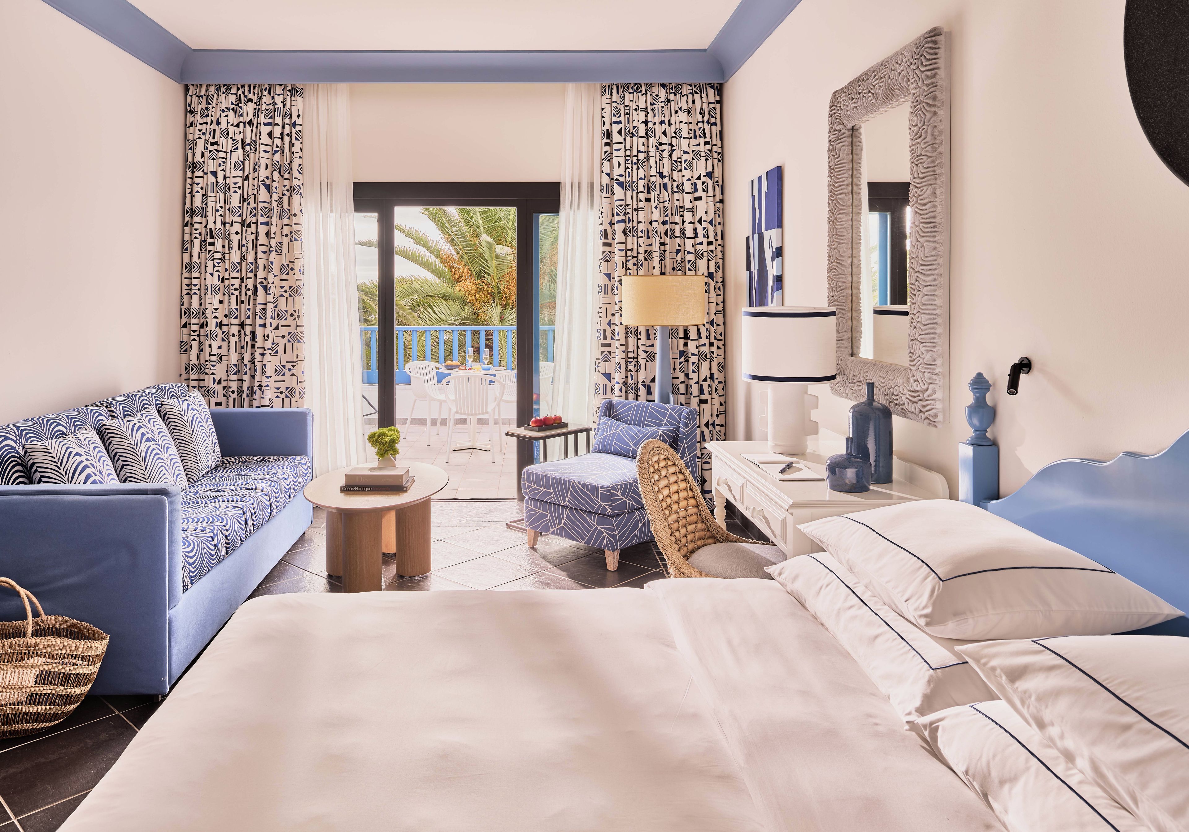 A hotel room with a bed, a sofa, a chair, a small table, and a balcony with a view of palm trees.