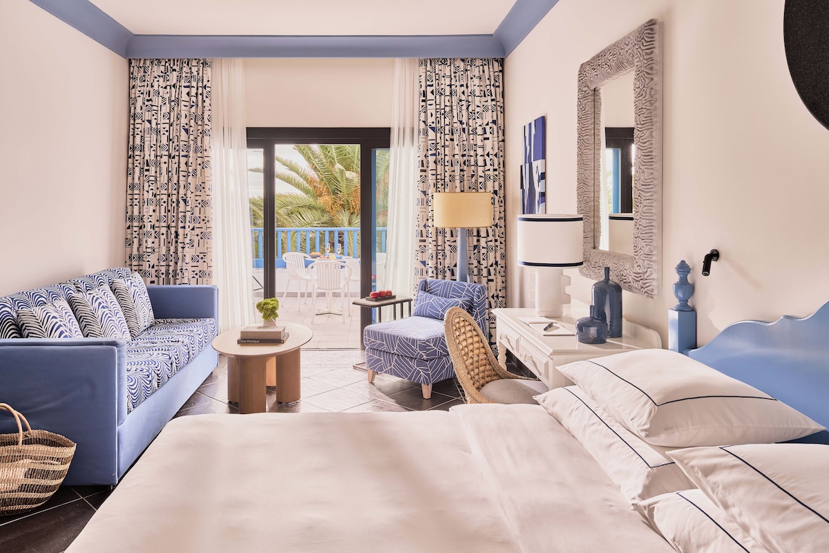 Habitación de hotel con cama, sofá, sillón, mesita pequeña y balcón con vistas a palmeras.