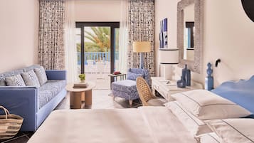 A hotel room with a bed, a sofa, a chair, a small table, and a balcony with a view of palm trees.