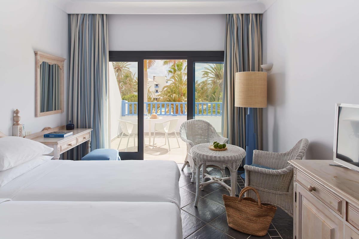 A hotel room with two beds, a desk, a chair, a small table with a fruit bowl, a TV, and a balcony with chairs and palm trees.