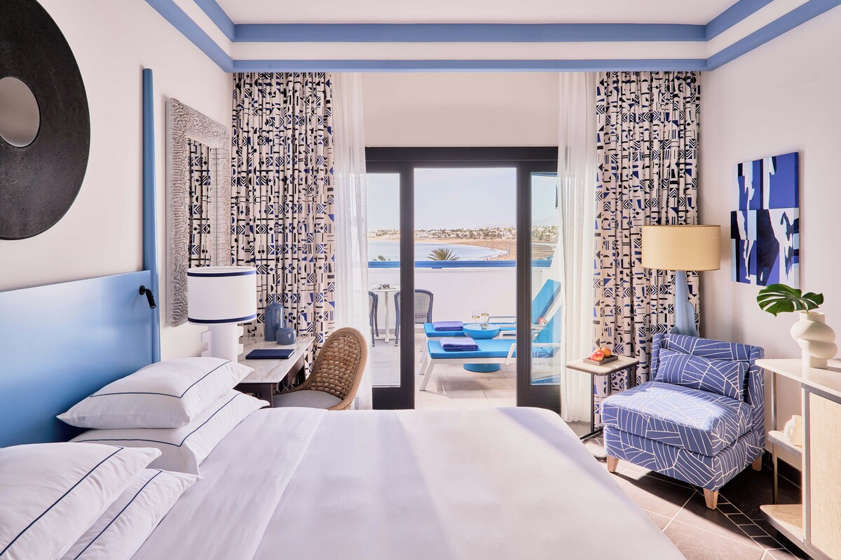 Habitación de hotel con cama, una silla, una mesa, una lámpara y vista a una piscina y playa.