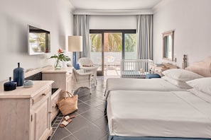 A hotel room with two beds, a desk, a chair, a crib, and a balcony with a view of palm trees.