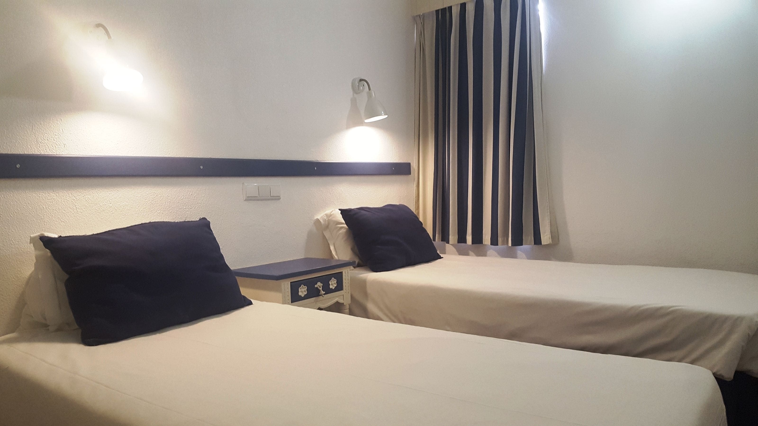 Hotellihuone, jossa on kaksi erillistä sänkyä, yöpöytä, lamppu ja ikkuna, jossa on raidalliset verhot.