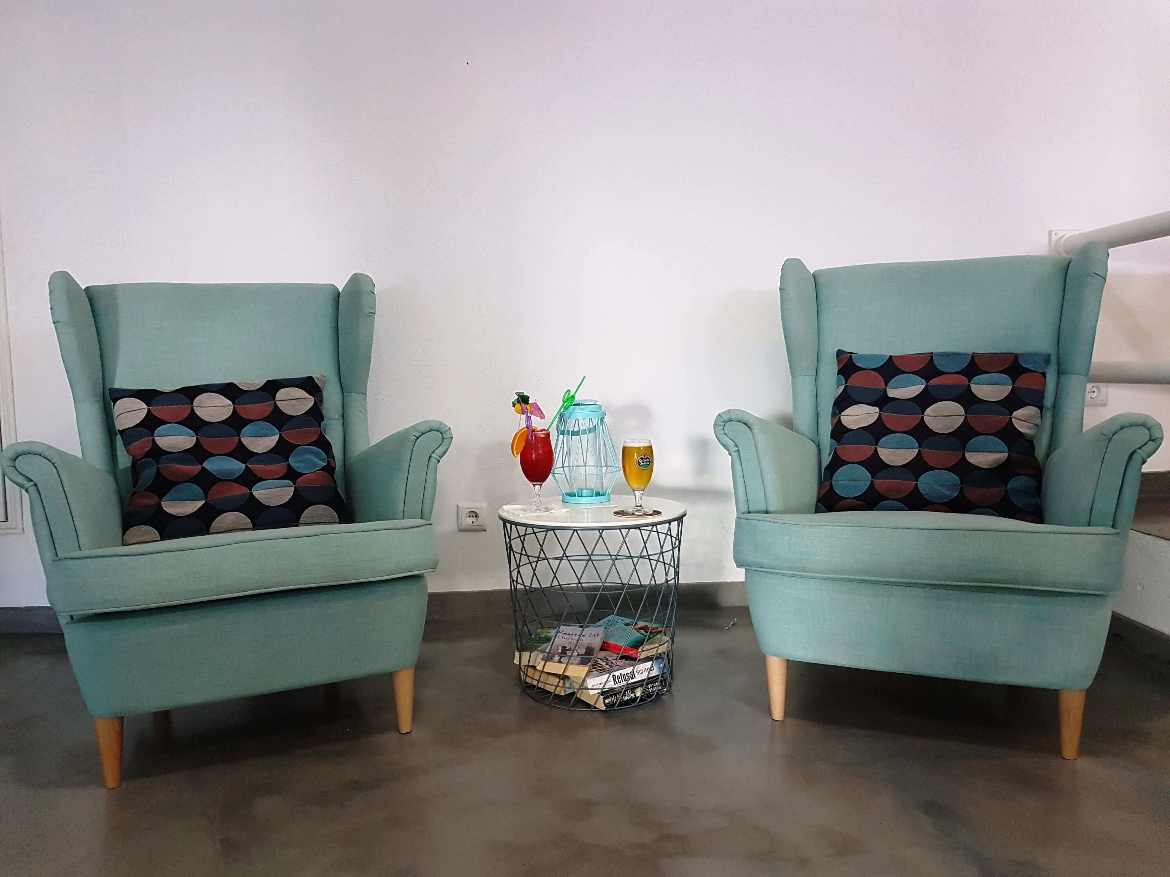 Two teal armchairs with patterned cushions, a small round table with drinks and a basket of magazines, and a plain white wall in the background.