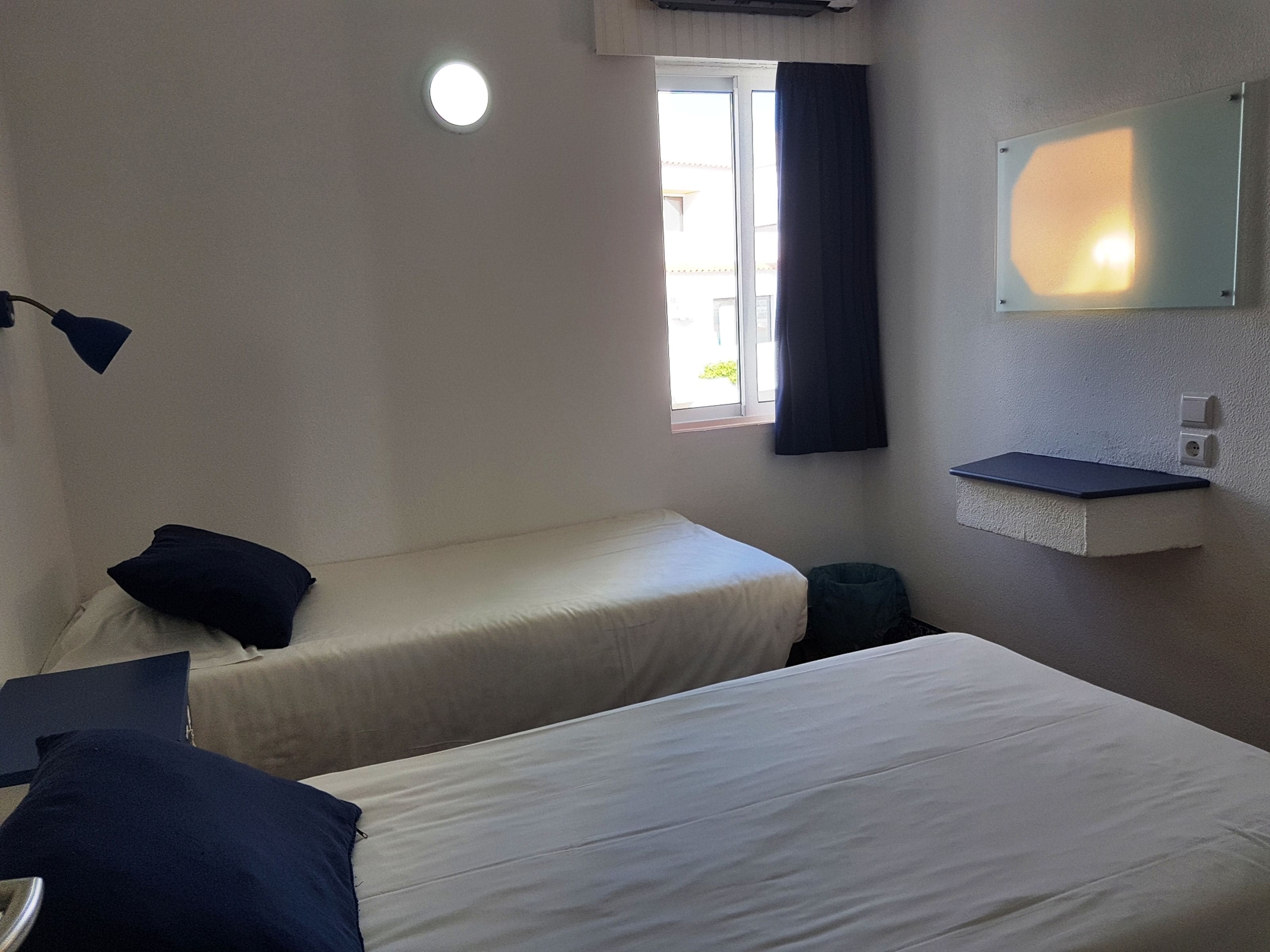 Una habitación con dos camas, una ventana pequeña, un espejo y una mesita de noche.