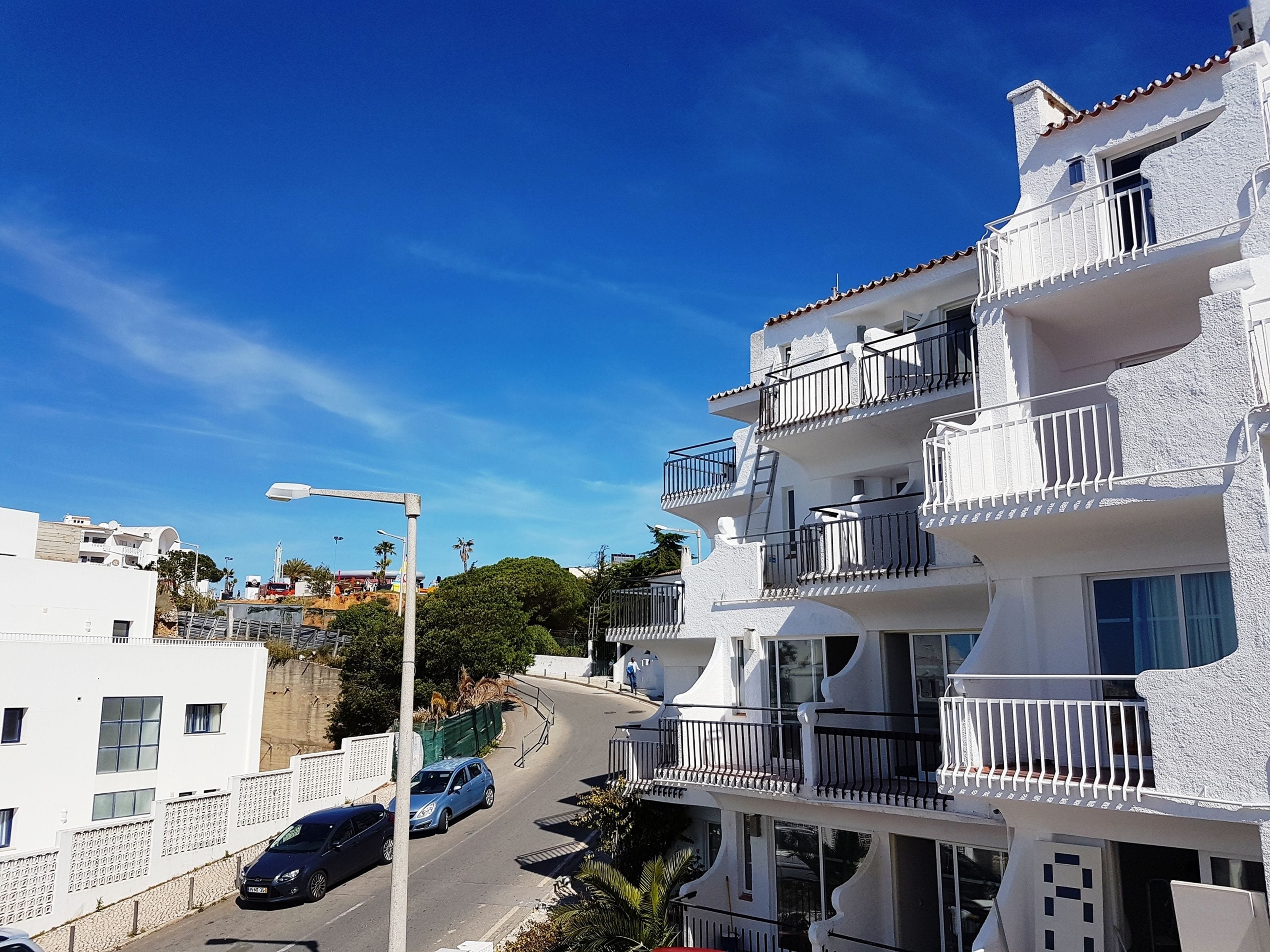 Un edificio blanco de varios pisos con balcones, una calle curva y coches aparcados.