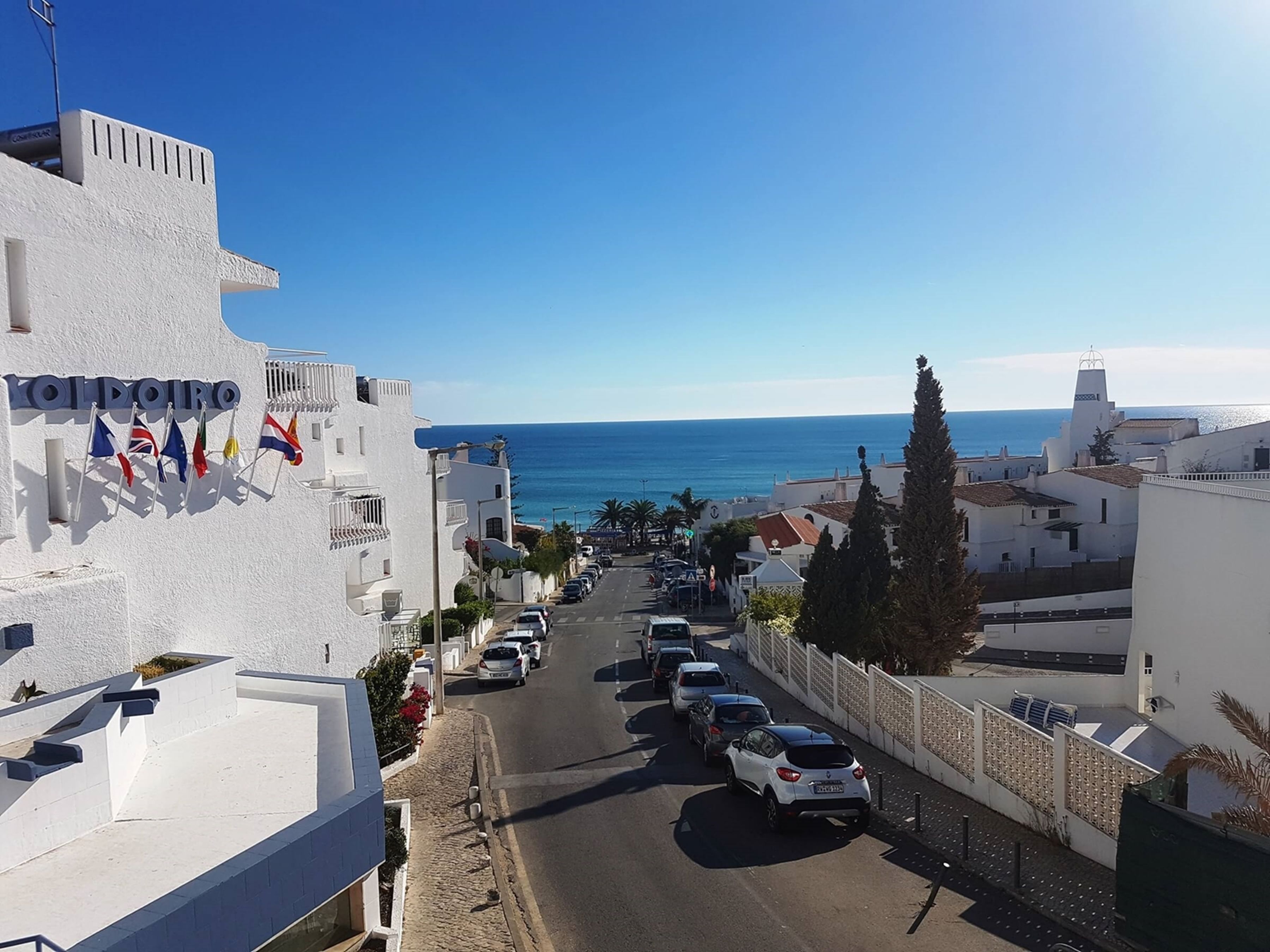 A coastal street with white buildings, a clear blue sky, and the sea in the background.