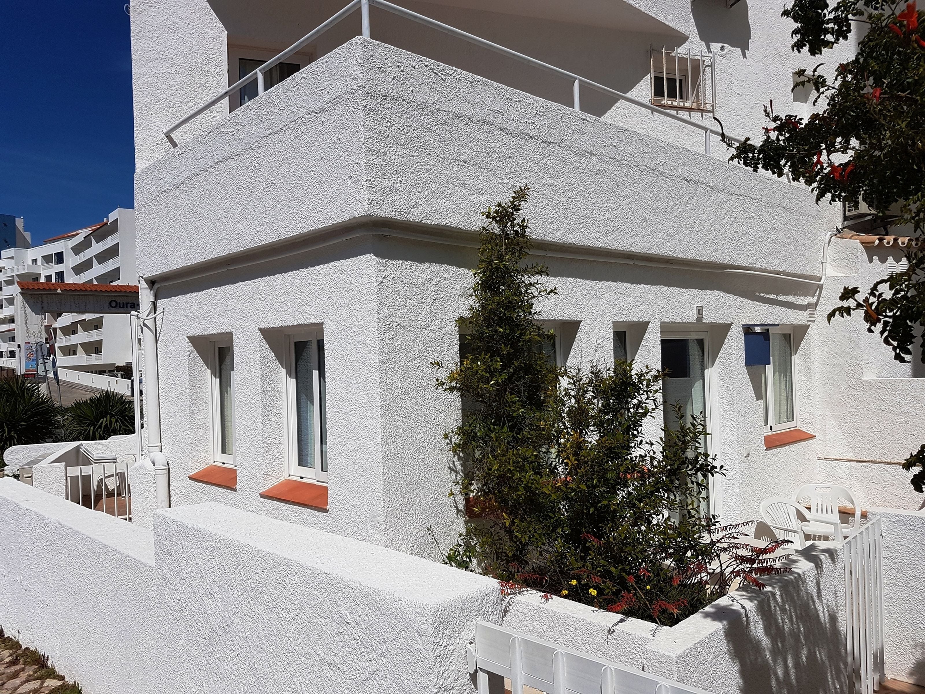 A white building with multiple windows and balconies, surrounded by greenery and other buildings.