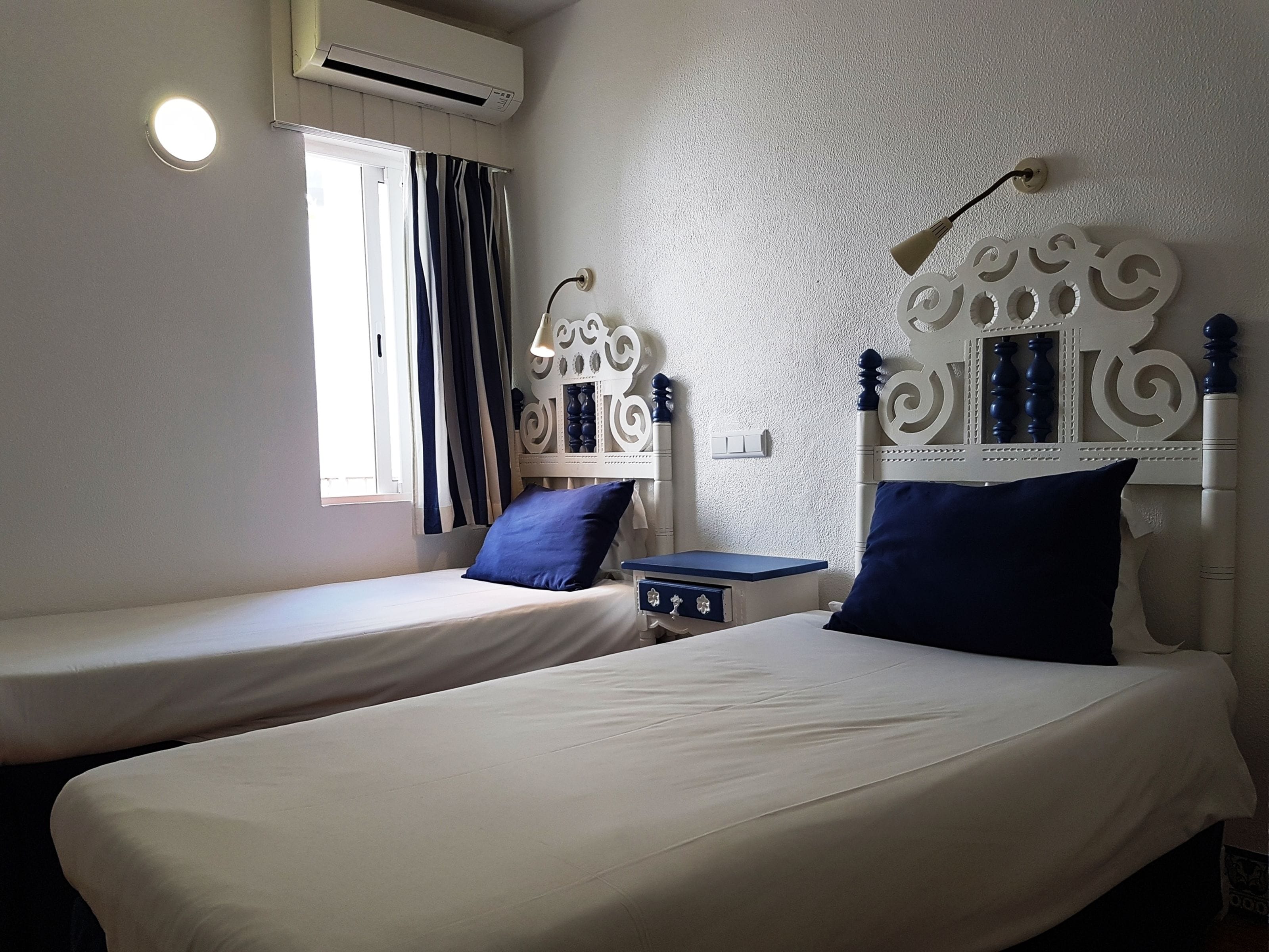 A room with two beds, each with a headboard and a pillow. There is a window with curtains, an air conditioning unit, and a wall-mounted light fixture.