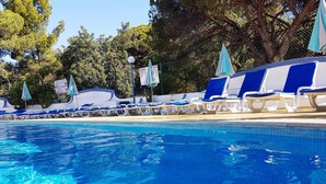 A swimming pool with blue water, surrounded by lounge chairs and umbrellas, with trees and a fence in the background. - Apartamentos Soldoiro (Albufeira)