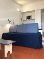 Espaço compacto com sofá, uma pequena mesa, uma luminária e área de kitchenette.