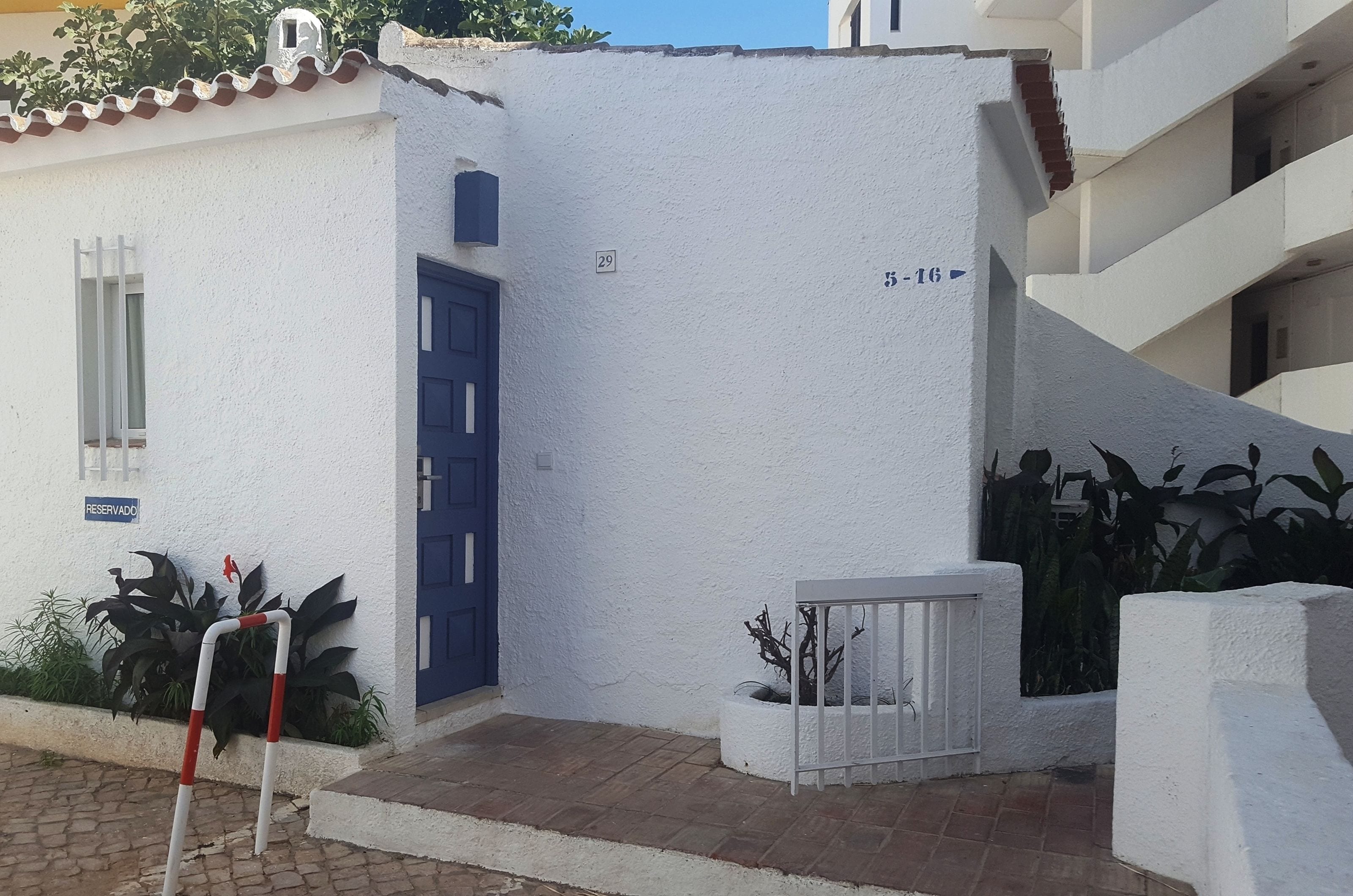 A white building with a blue door, numbered 29, and a small garden area with plants.