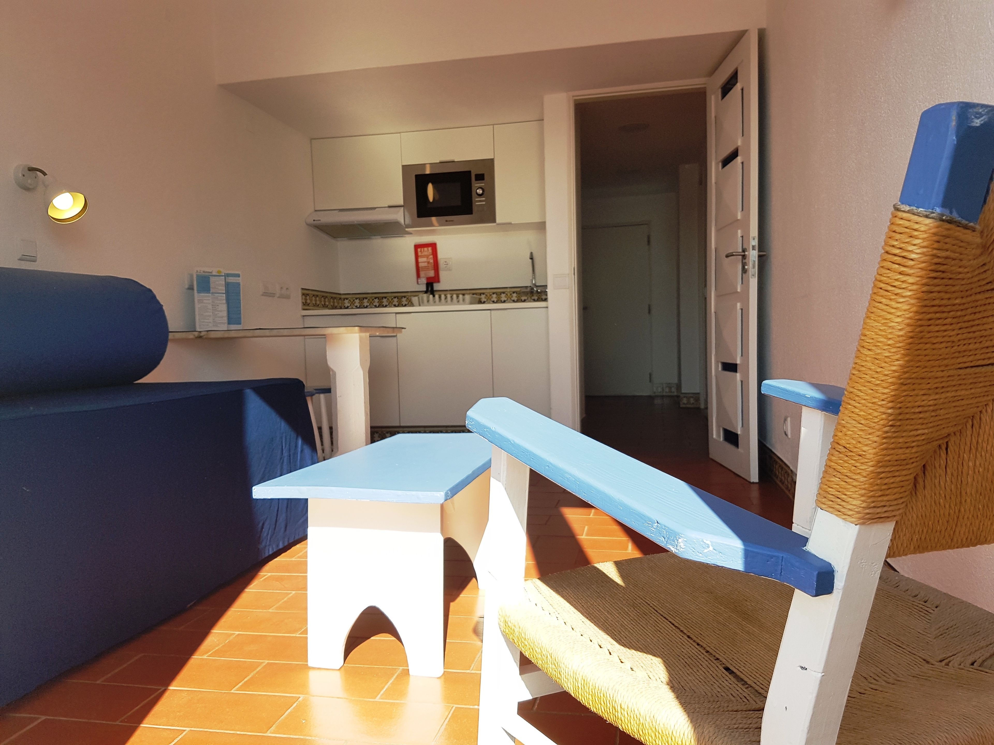Ein gemütlicher, kleiner Wohnbereich mit Küchenzeile, einem blauen Sofa, einem kleinen Tisch und einem Korbstuhl.