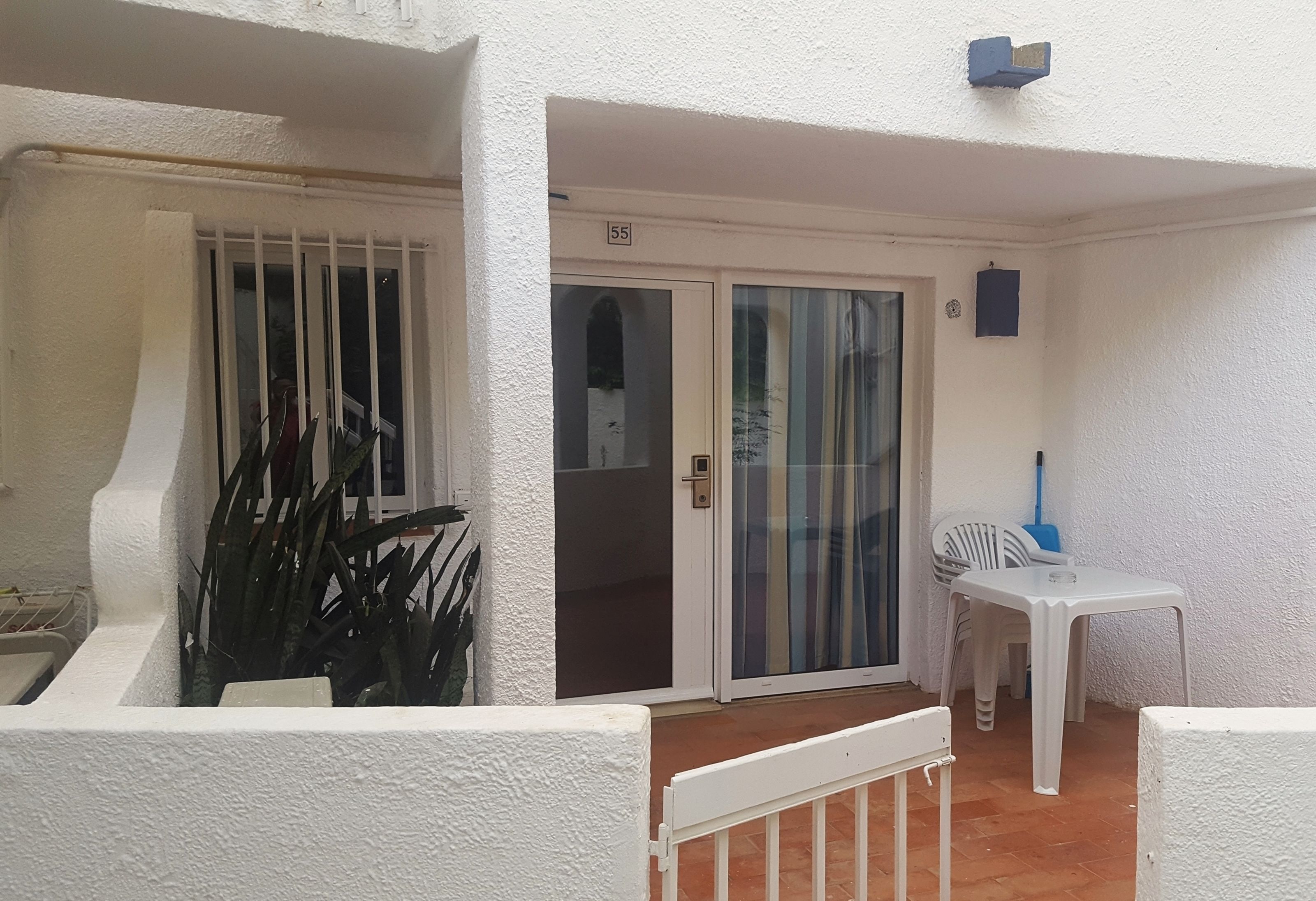 Un balcon blanc avec une porte-fenêtre coulissante, une petite table avec des chaises et un balai appuyé contre le mur.