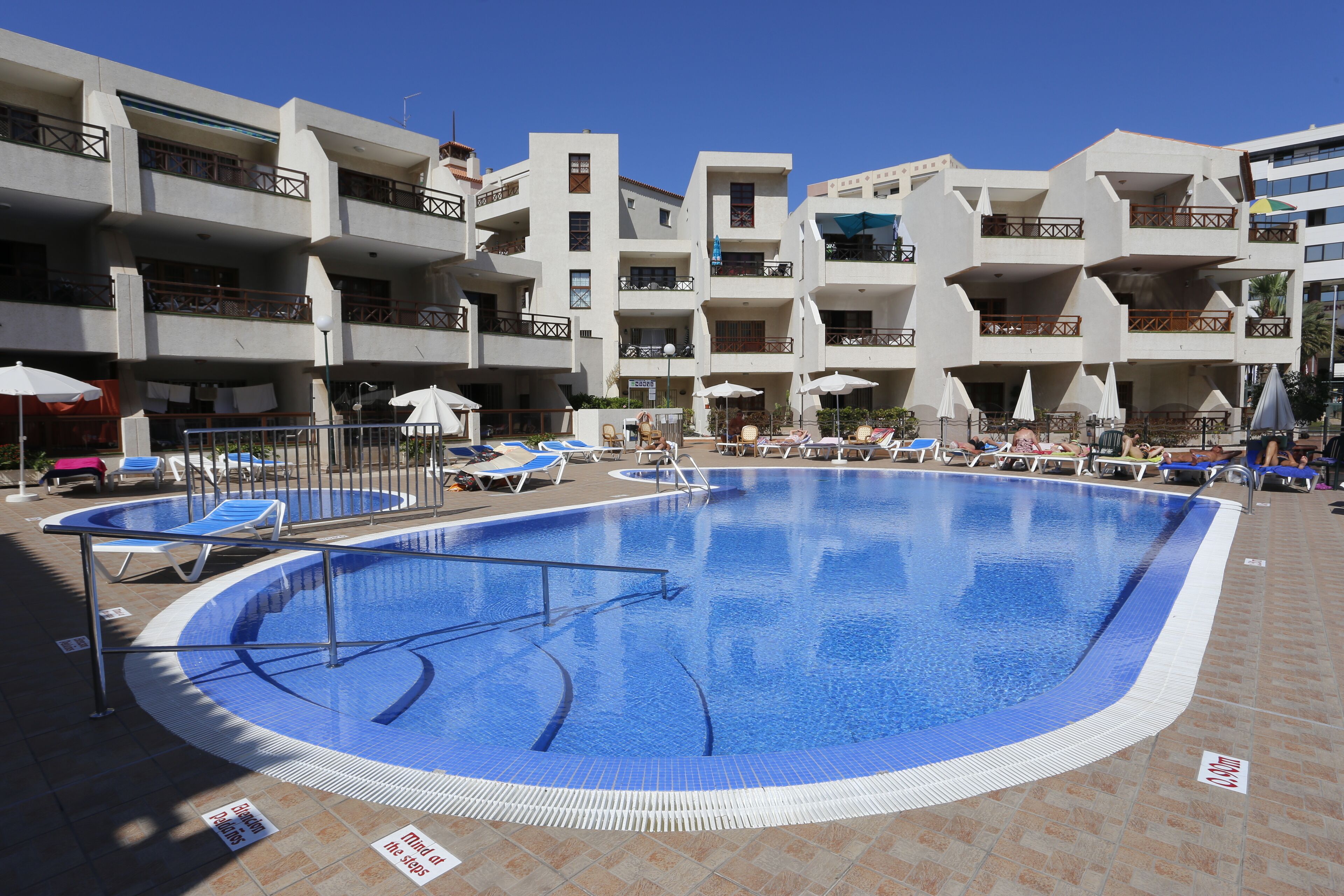 Una piscina con sillas de descanso y sombrillas, rodeada de edificios con balcones.