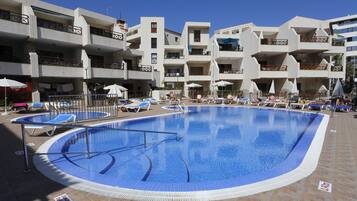 Una piscina con sillas de descanso y sombrillas, rodeada de edificios con balcones.