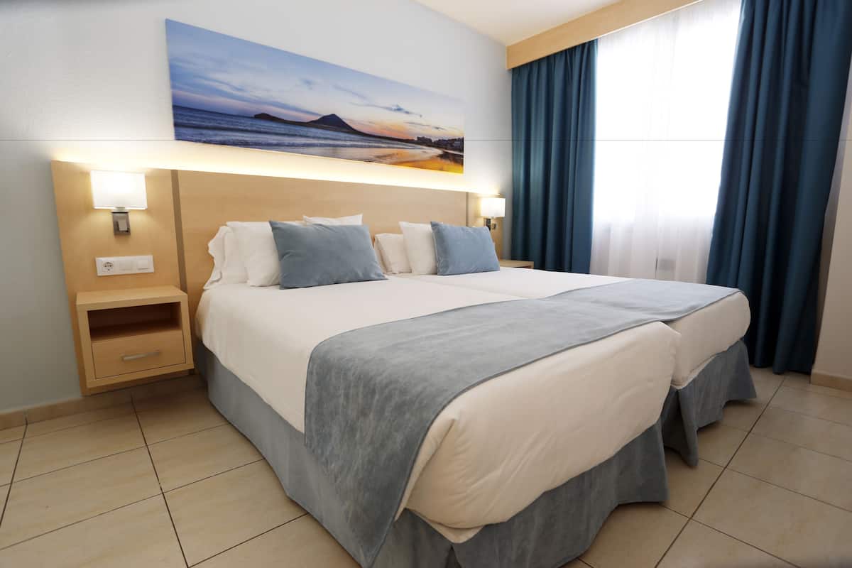 A double bed with a grey bed skirt, two bedside tables with lamps, a nightstand, and a large window with blue curtains.