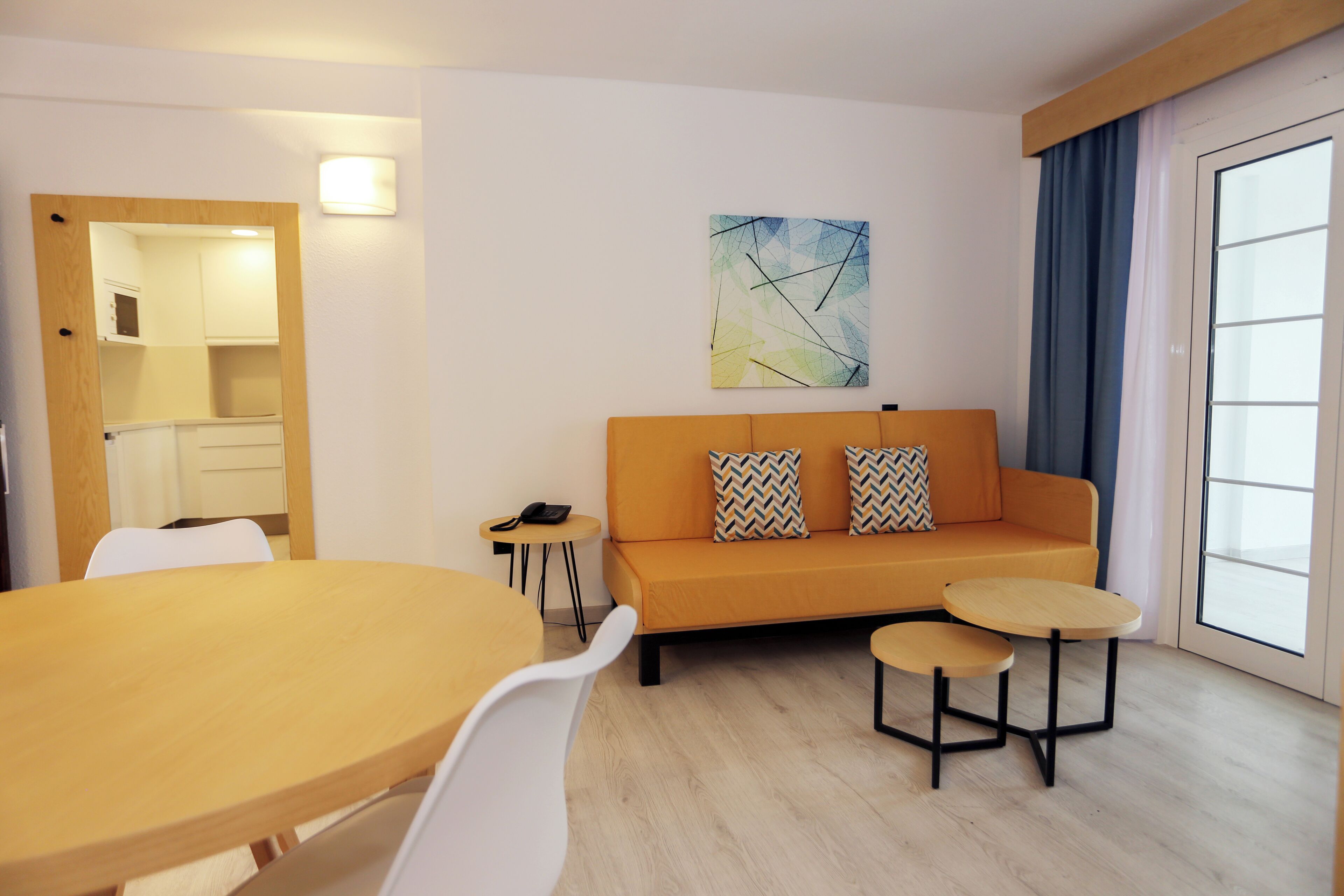 A modern living room with a sofa, two small tables, a dining table, and a kitchen area visible through an open door.