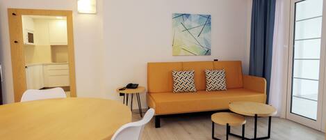 A modern living room with a sofa, two small tables, a dining table, and a kitchen area visible through an open door.