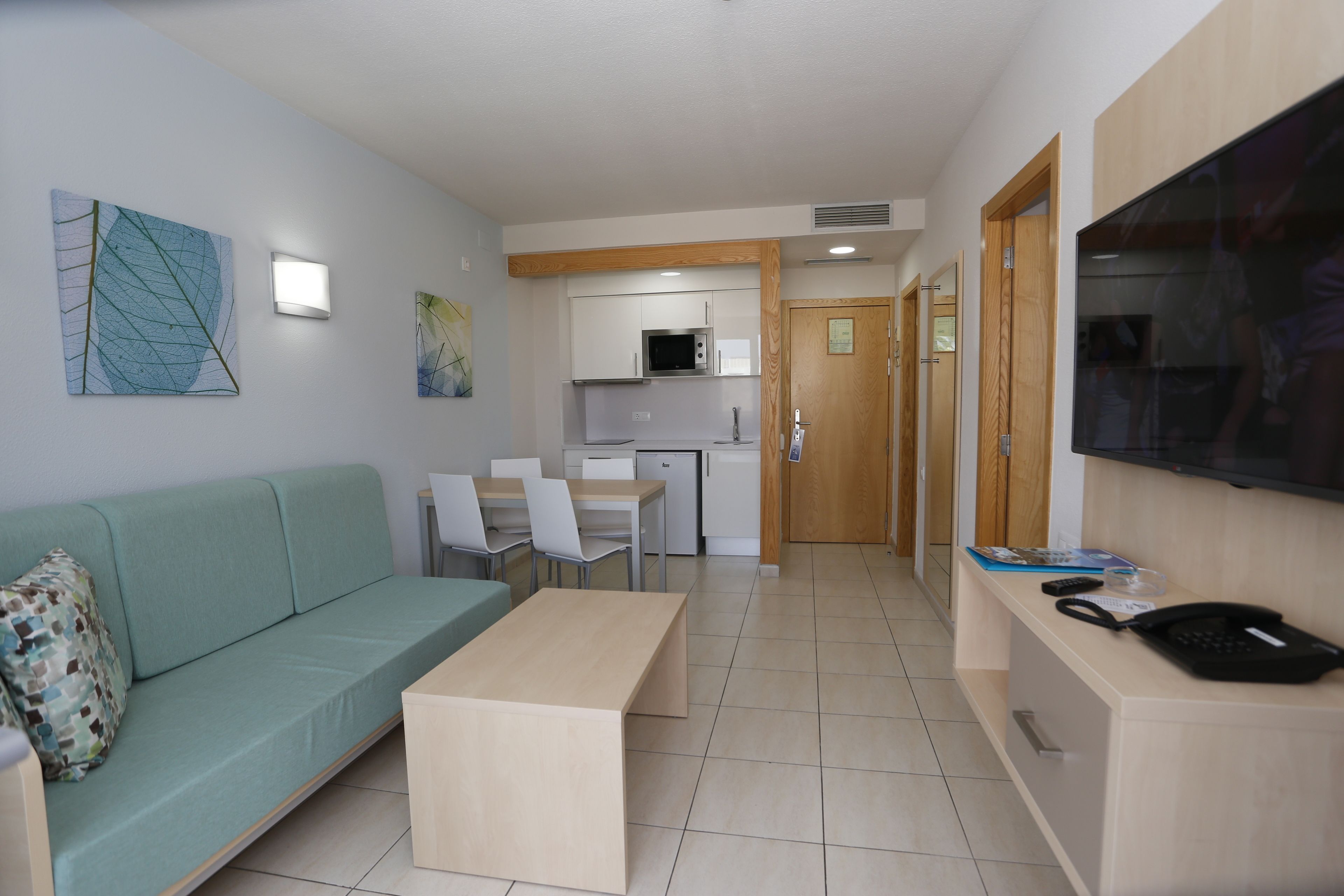 A modern living room with a sofa, a coffee table, and a TV. There is a kitchen area with a microwave and cabinets.