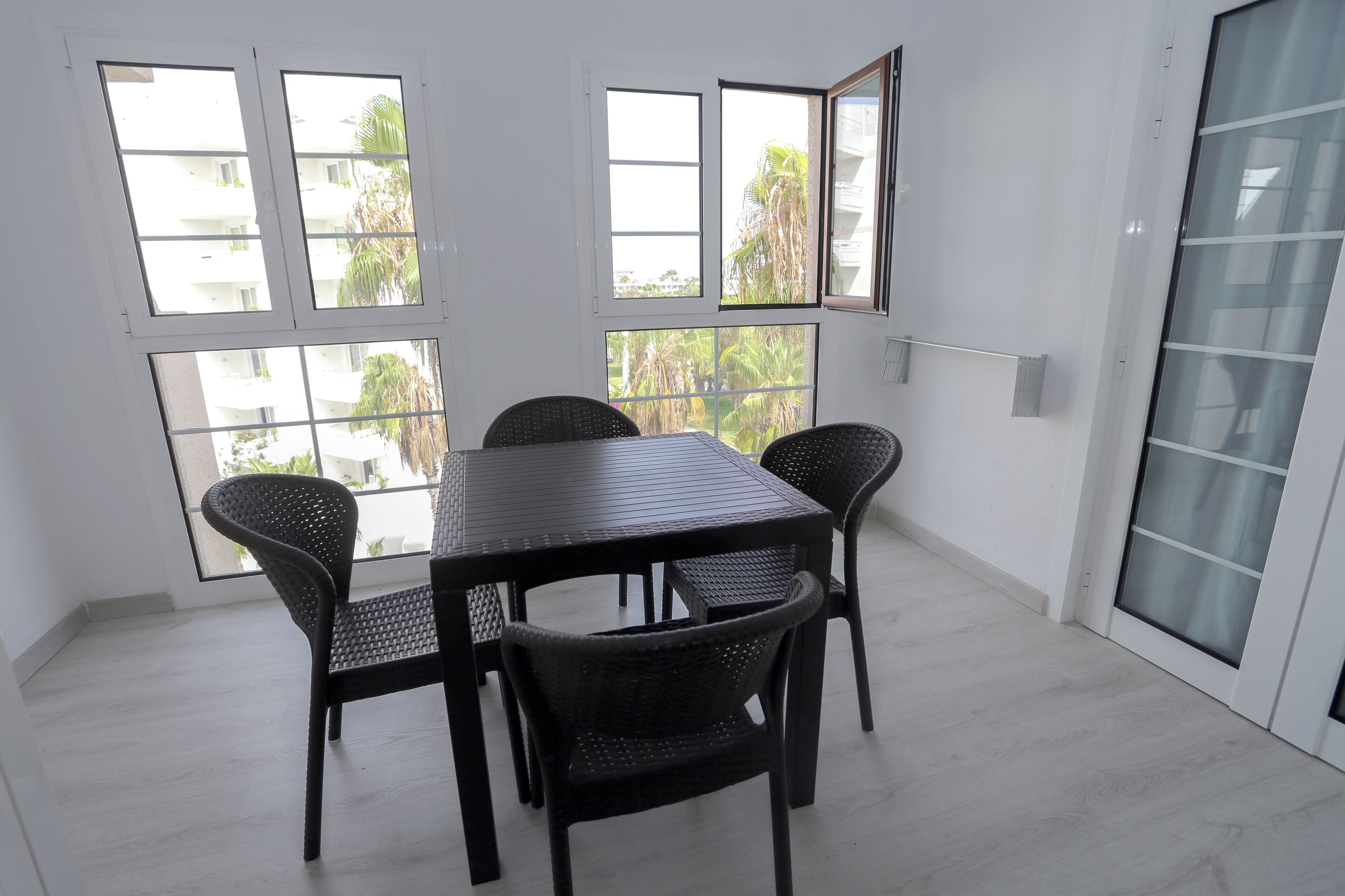 A room with a dining table and chairs, large windows with a view of palm trees, and a light-colored floor.