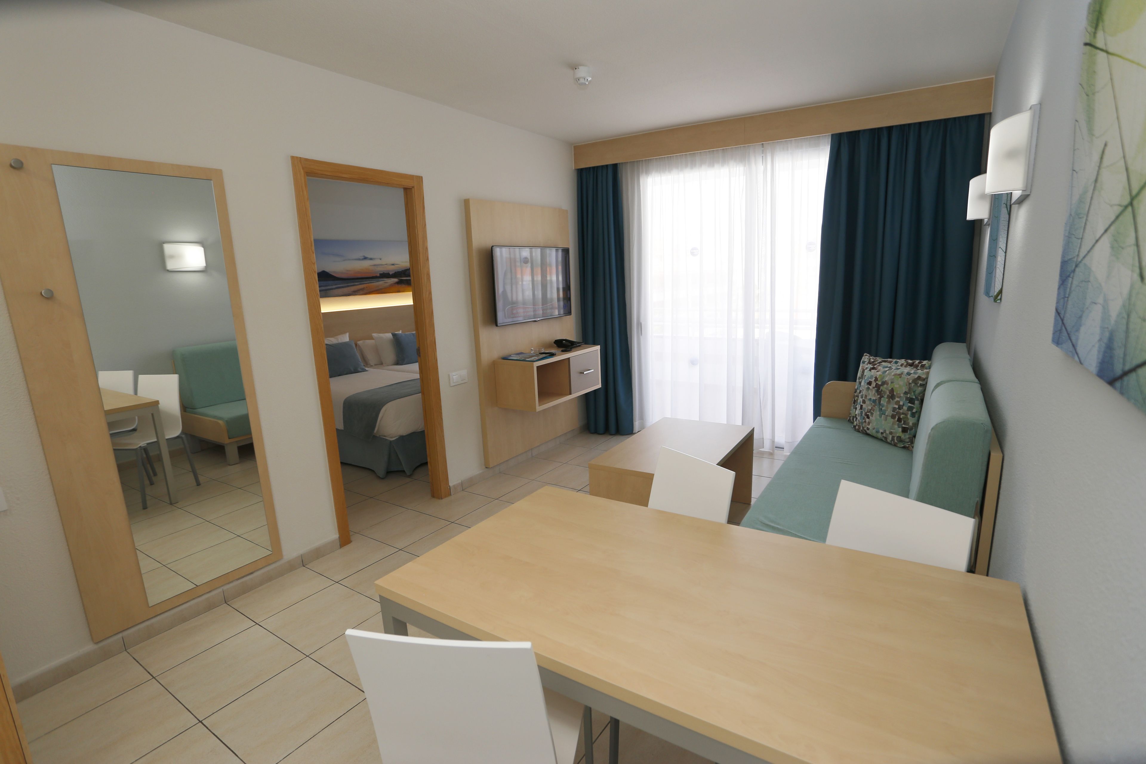 A modern hotel room with a dining area, a sofa, a TV, and a large window with curtains.