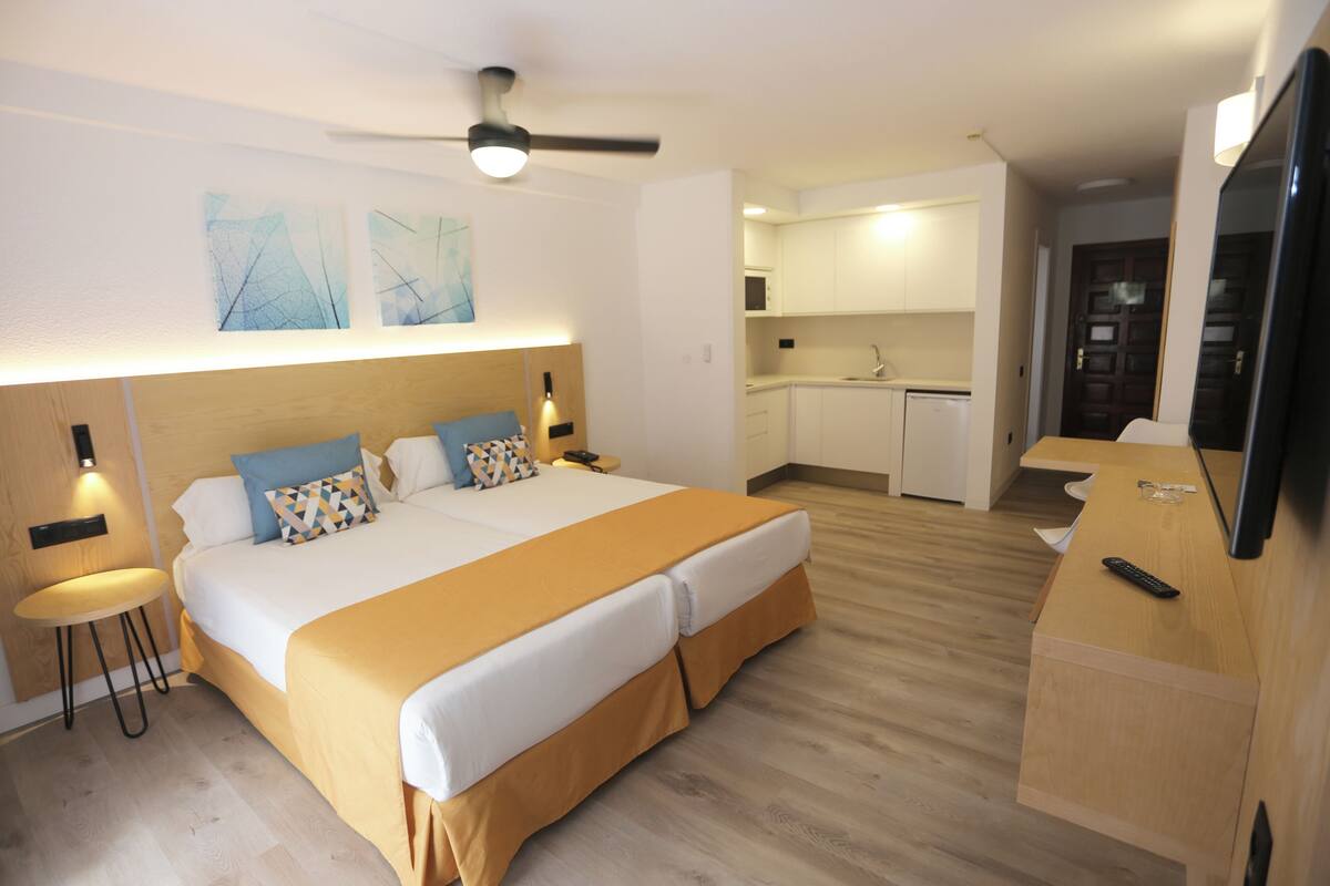 A modern hotel room with a large bed, a small kitchen area, and a TV.