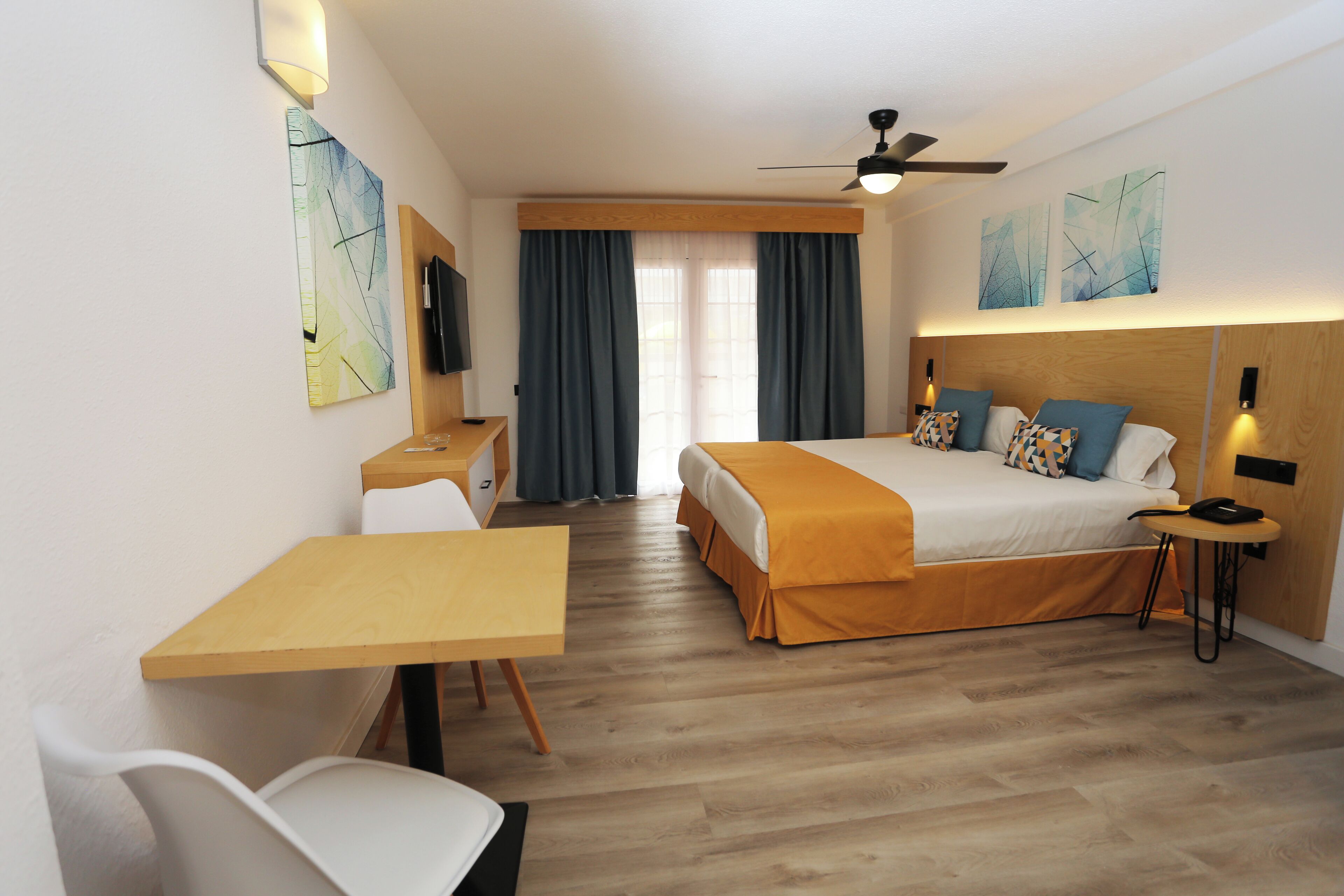 A hotel room with a bed, a desk, a chair, a TV, and a ceiling fan.