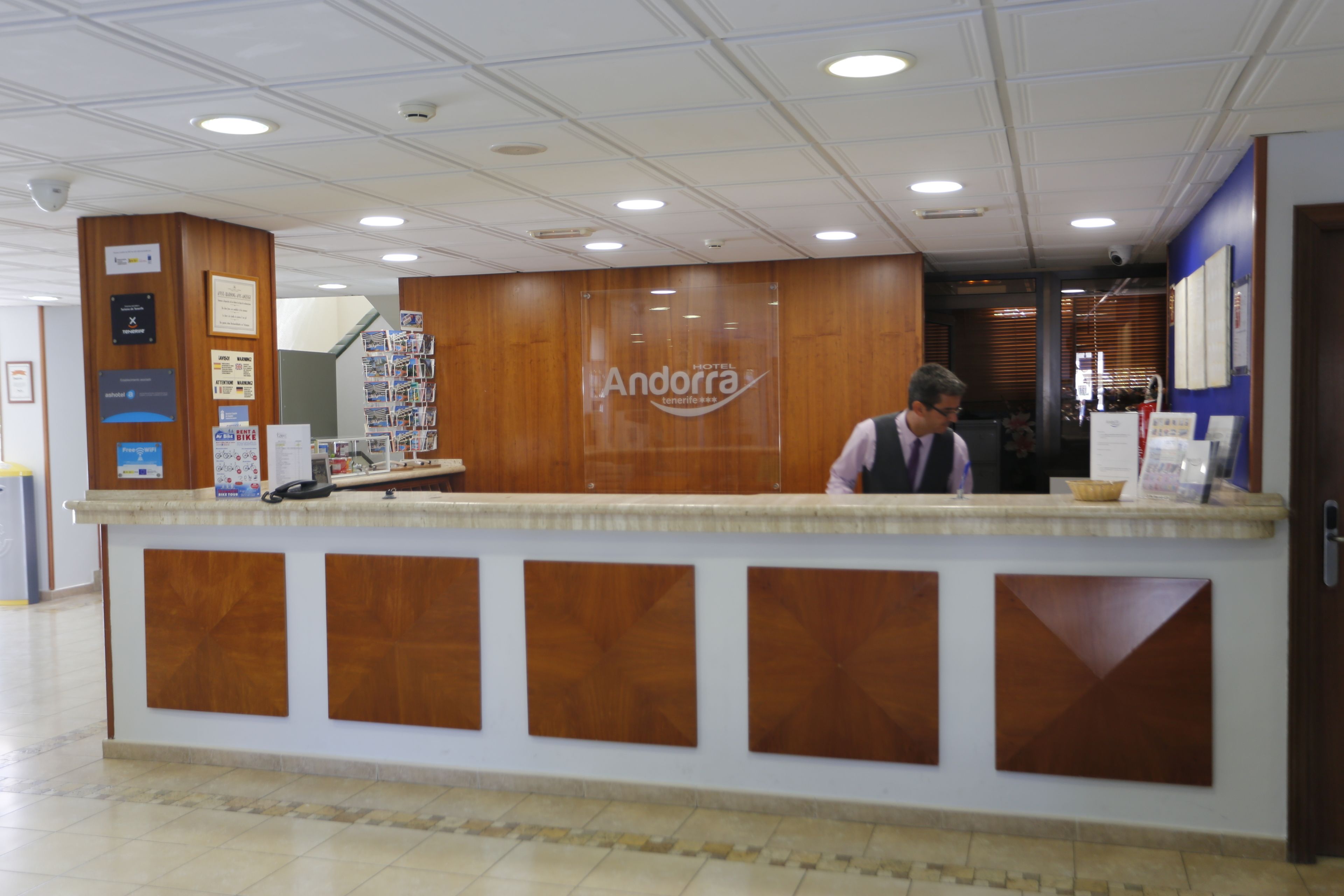 Un comptoir d’accueil d’hôtel avec un revêtement en bois, une enseigne indiquant « Andorre » et un présentoir avec divers articles.