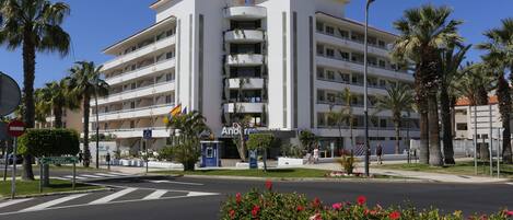 Un hotel de varios pisos con balcones, una calle al frente y palmeras.