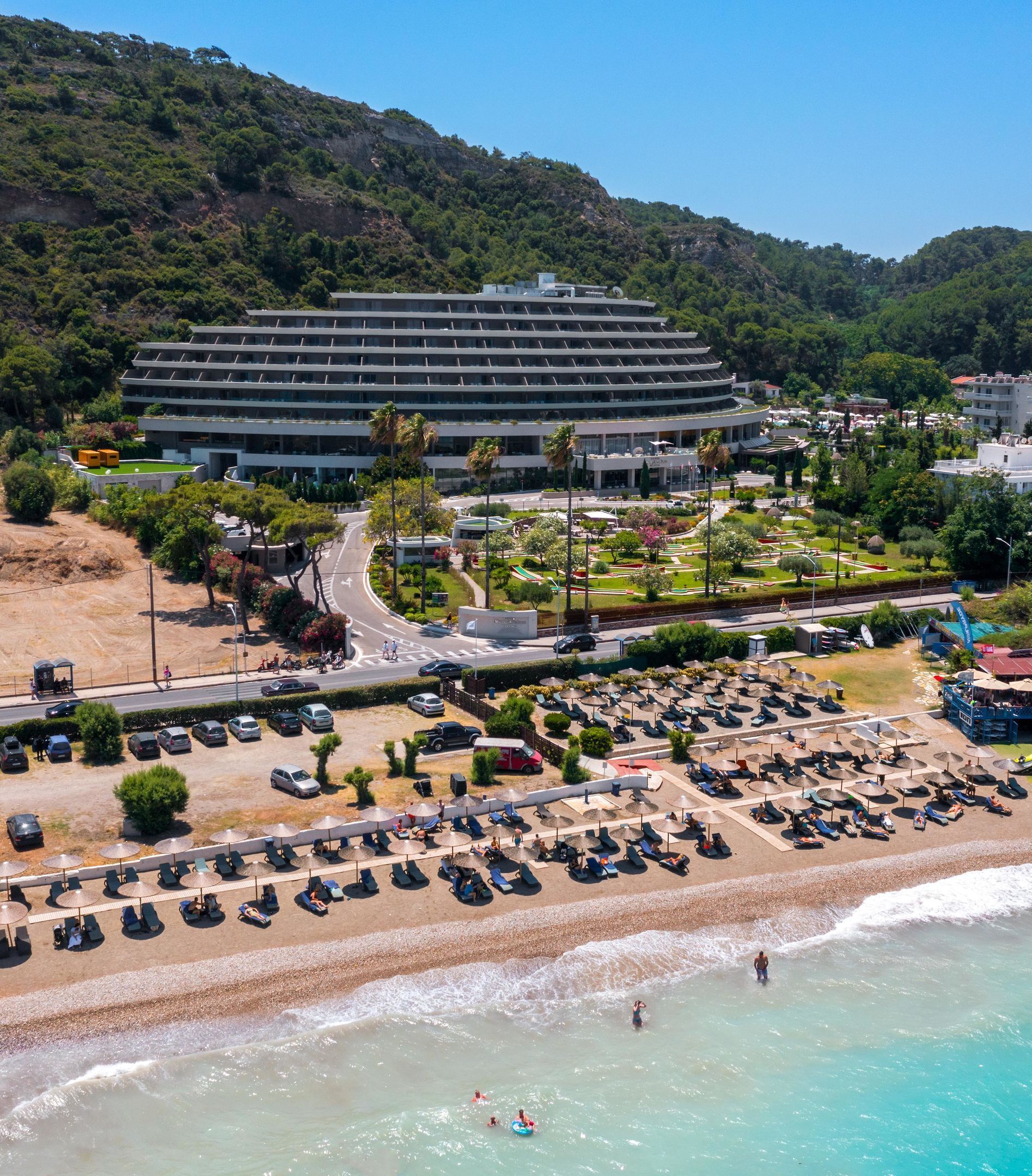 Un hotel costero con un edificio grande, un estacionamiento y una playa con tumbonas.