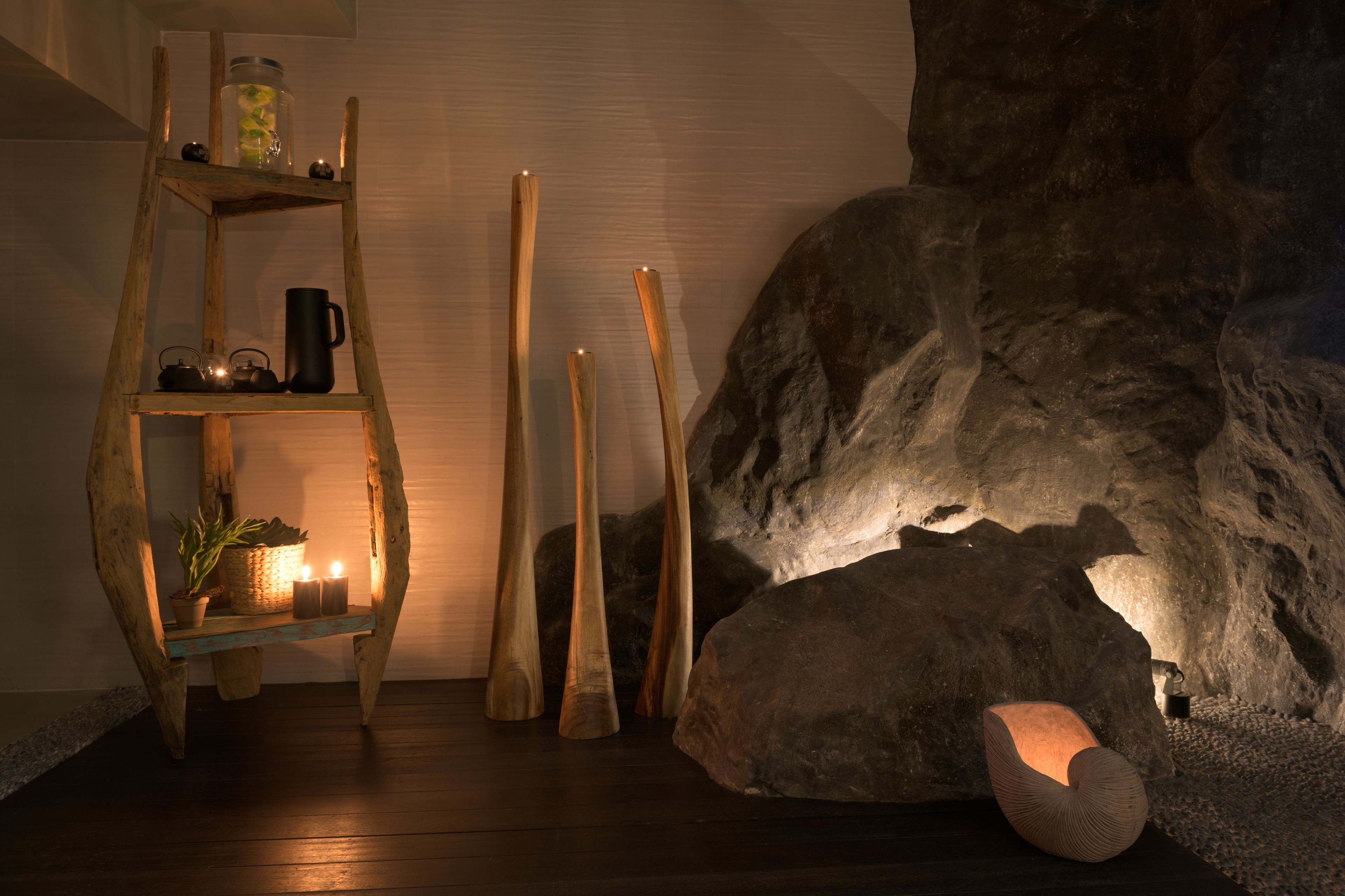 Een rustiek interieur met een houten laddenplank waarop een potplant, een zwarte mok en een glazen pot staan. Een grote rotsformatie wordt verlicht door een warm licht, en er is een unieke, gebogen zithoek te zien.