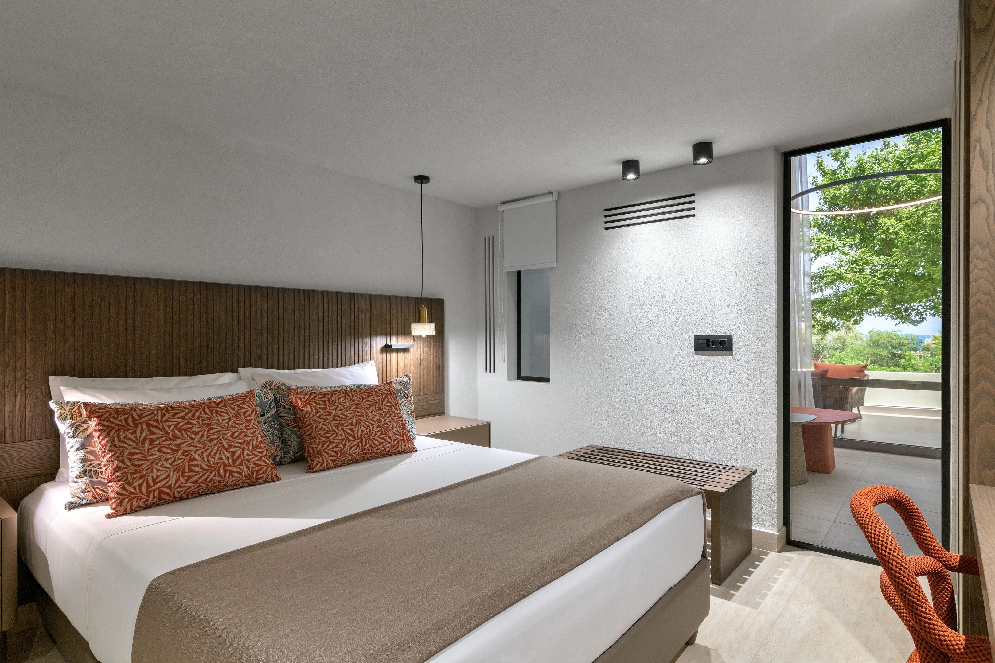 Una habitación de hotel moderna con una cama grande, cabecera de madera, mesita de noche, televisor montado en la pared y una puerta corrediza de cristal que da a un patio exterior.