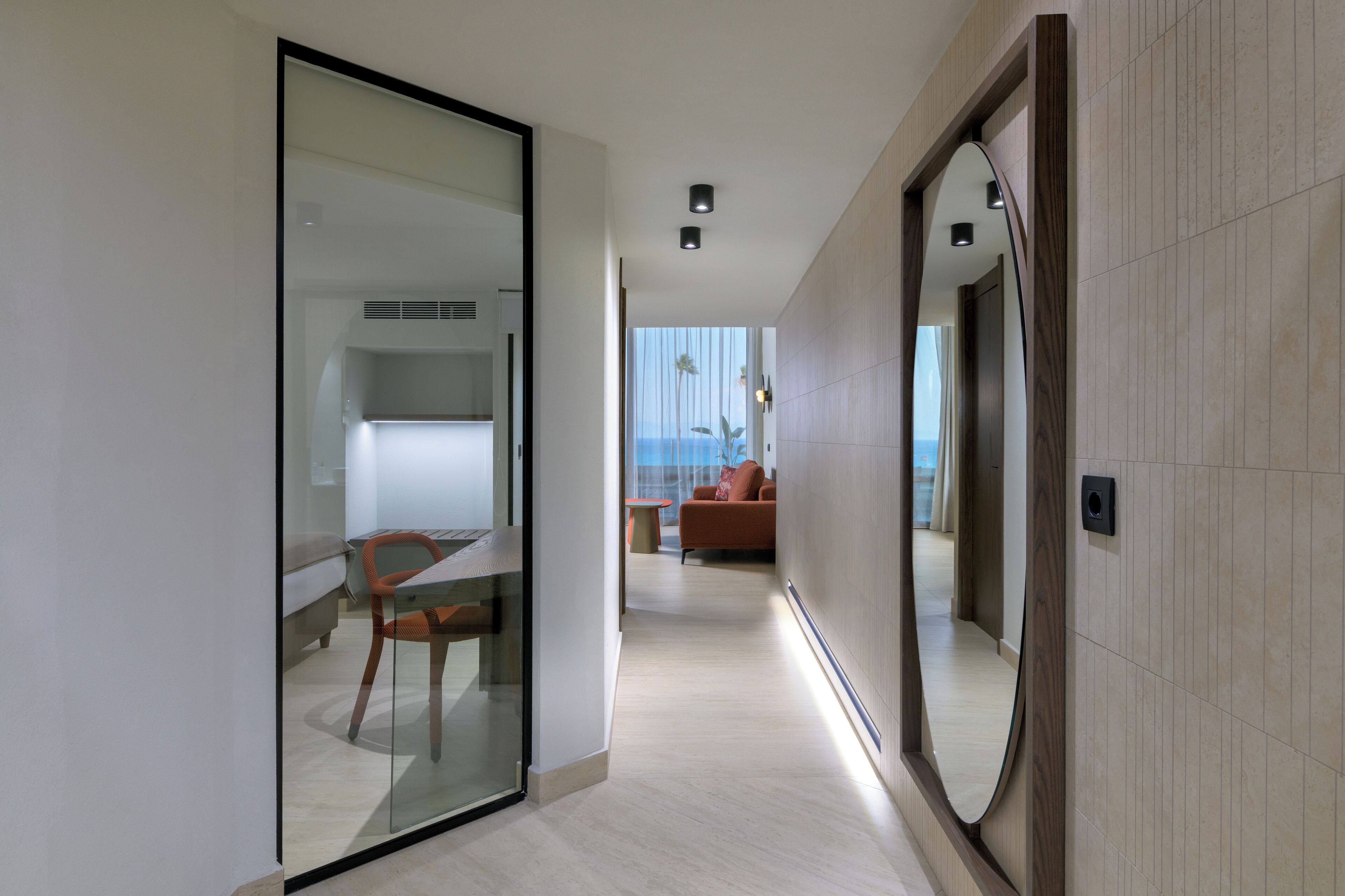 A modern hotel corridor with a glass partition, a wooden-framed mirror, and a view of a living area with a sofa and a table.