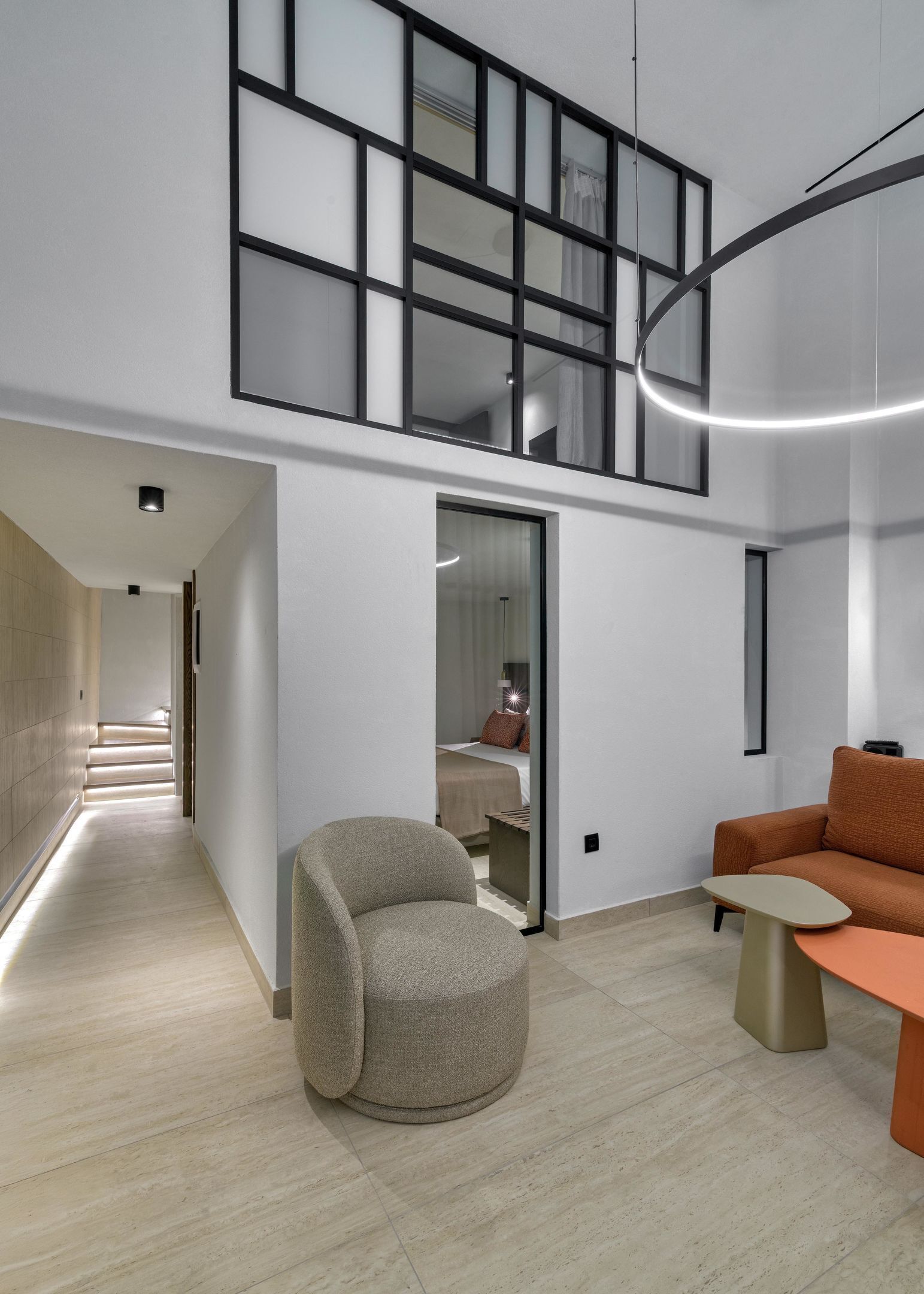 A modern interior with a high ceiling, a staircase leading to an upper level, a seating area with a sofa and a small table, and a bed visible through a glass door.