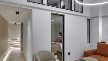 A modern interior with a high ceiling, a staircase leading to an upper level, a seating area with a sofa and a small table, and a bed visible through a glass door.
