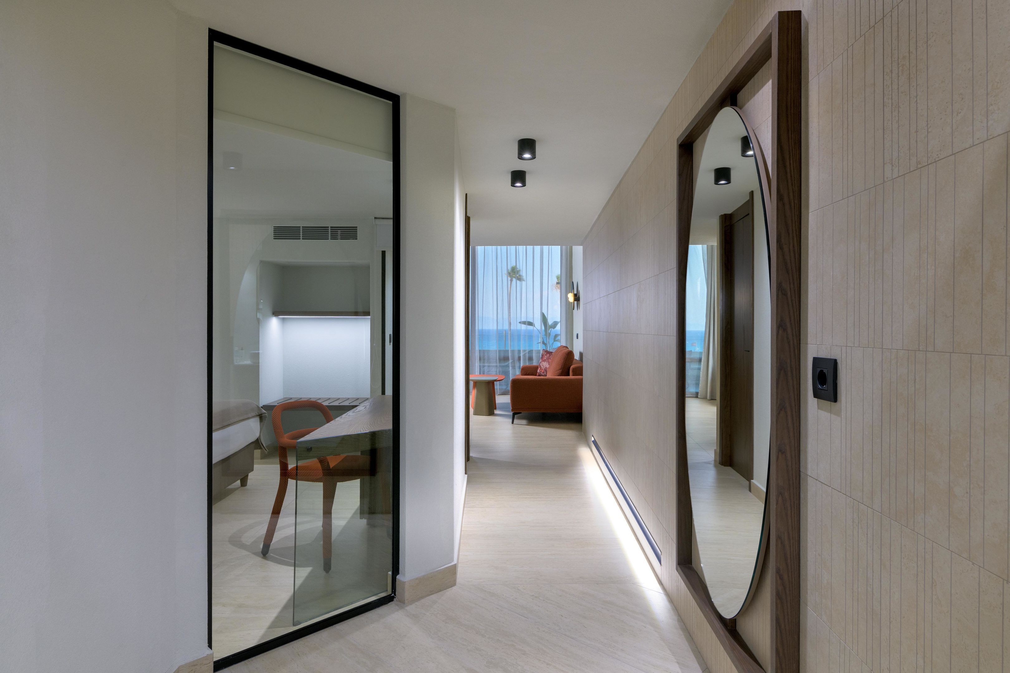 Un pasillo moderno de hotel con un panel de vidrio, un espejo con marco de madera y vistas a una sala de estar con sofá y mesa.