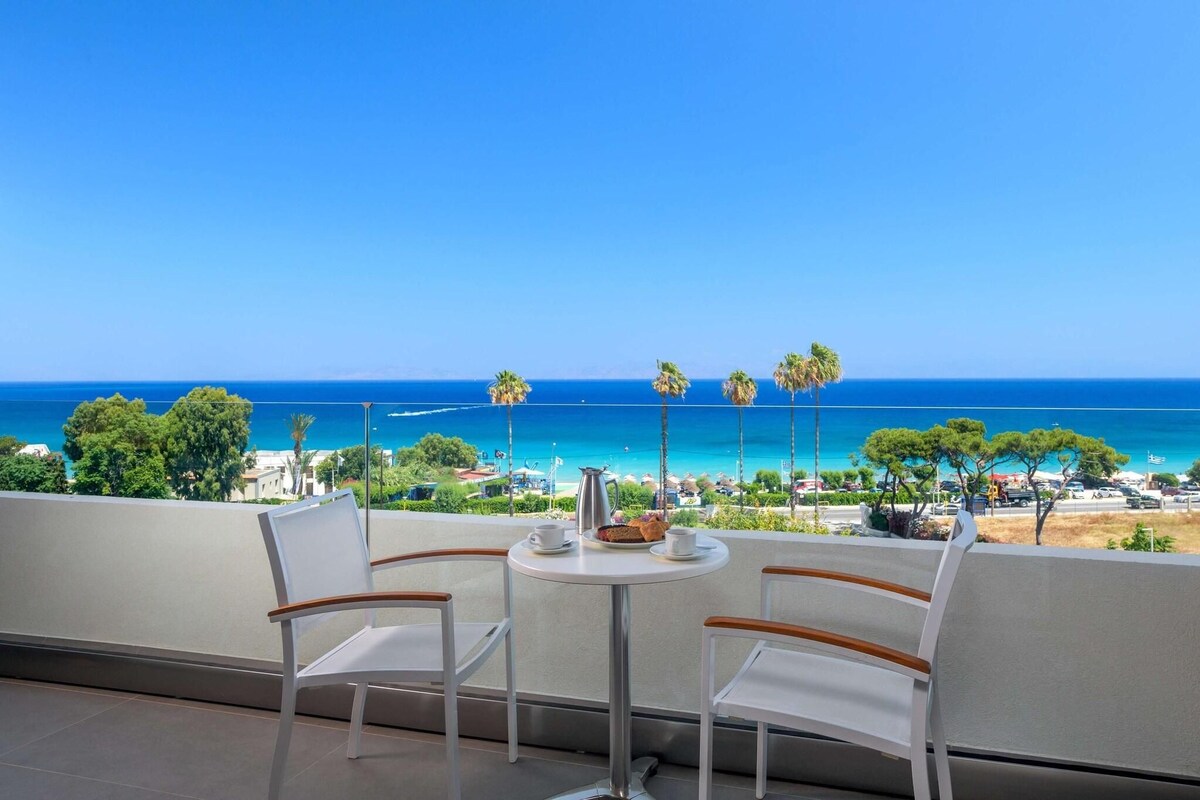 Un balcon avec deux chaises blanches et une petite table ronde, donnant sur une plage bordée de palmiers et sous un ciel bleu azur.