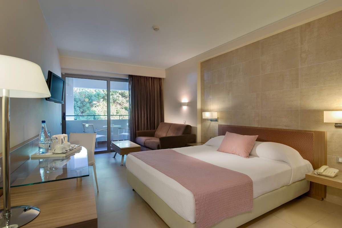 Une chambre d’hôtel comprenant un lit, un bureau avec une lampe, une chaise, une petite table et un téléviseur fixé au mur.