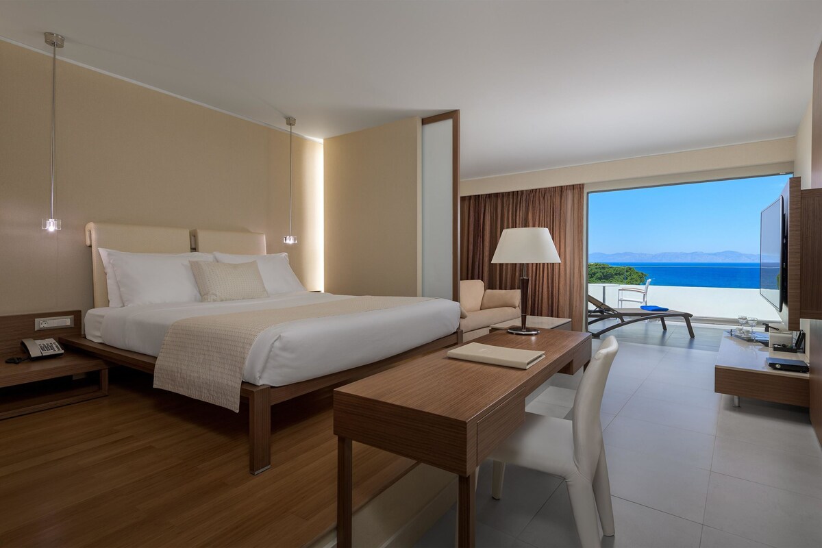 Een hotelkamer met een bed, bureau en uitzicht op zee.