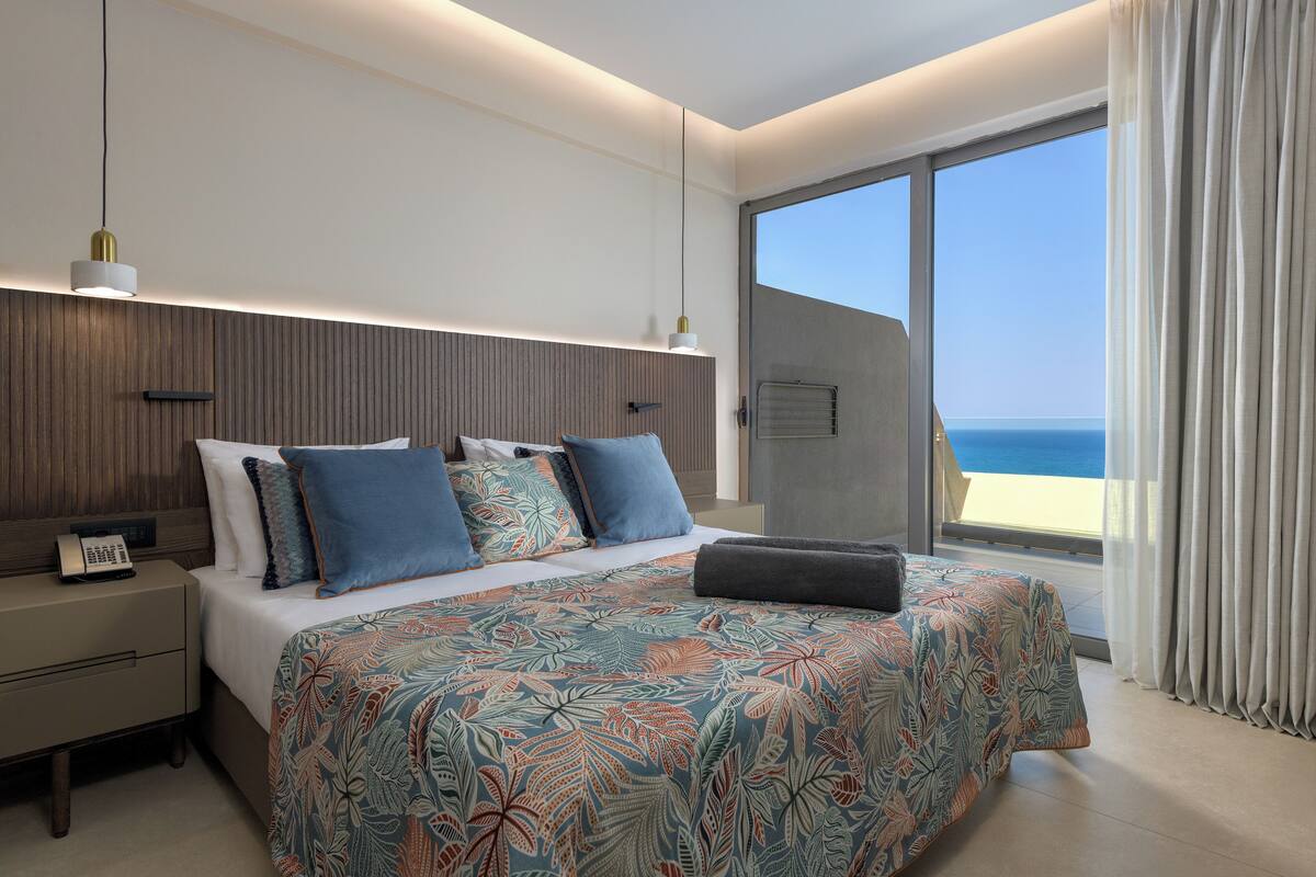 A modern hotel room with a large bed, a nightstand, a telephone, and a view of the sea through a sliding glass door.