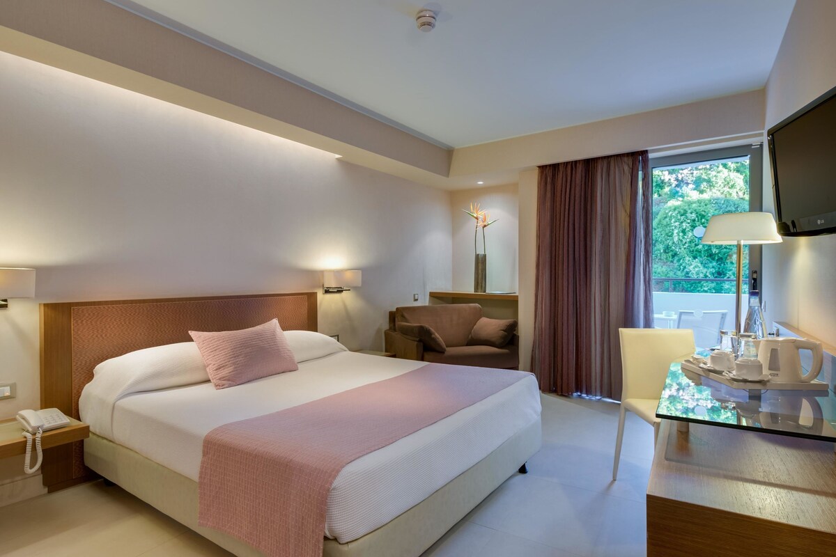 Een hotelkamer met een groot bed, nachtlampjes, een bureau met telefoon, een stoel, een tafel met een dienblad en een televisie.