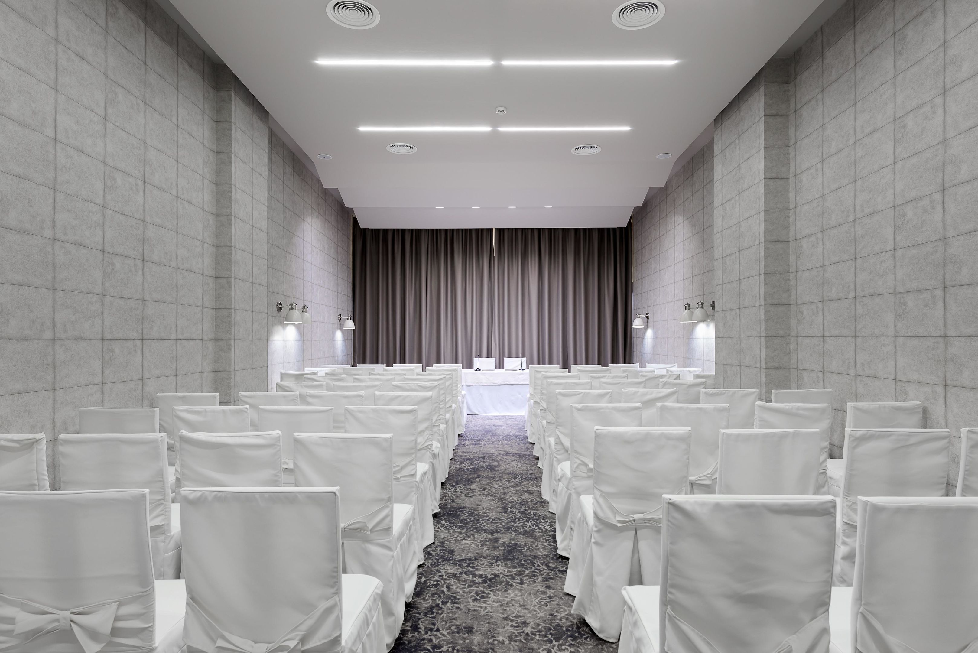 Un salón de conferencias con filas de sillas blancas, un escenario con una mesa blanca y un telón de fondo gris.