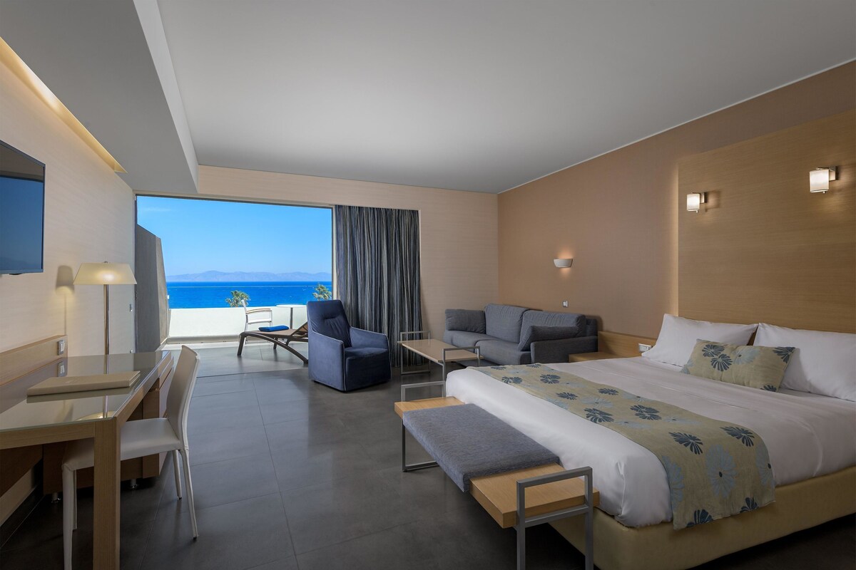 Een moderne hotelkamer met een groot bed, een bureau, een stoel en een bank. Er is een groot raam met uitzicht op de oceaan.