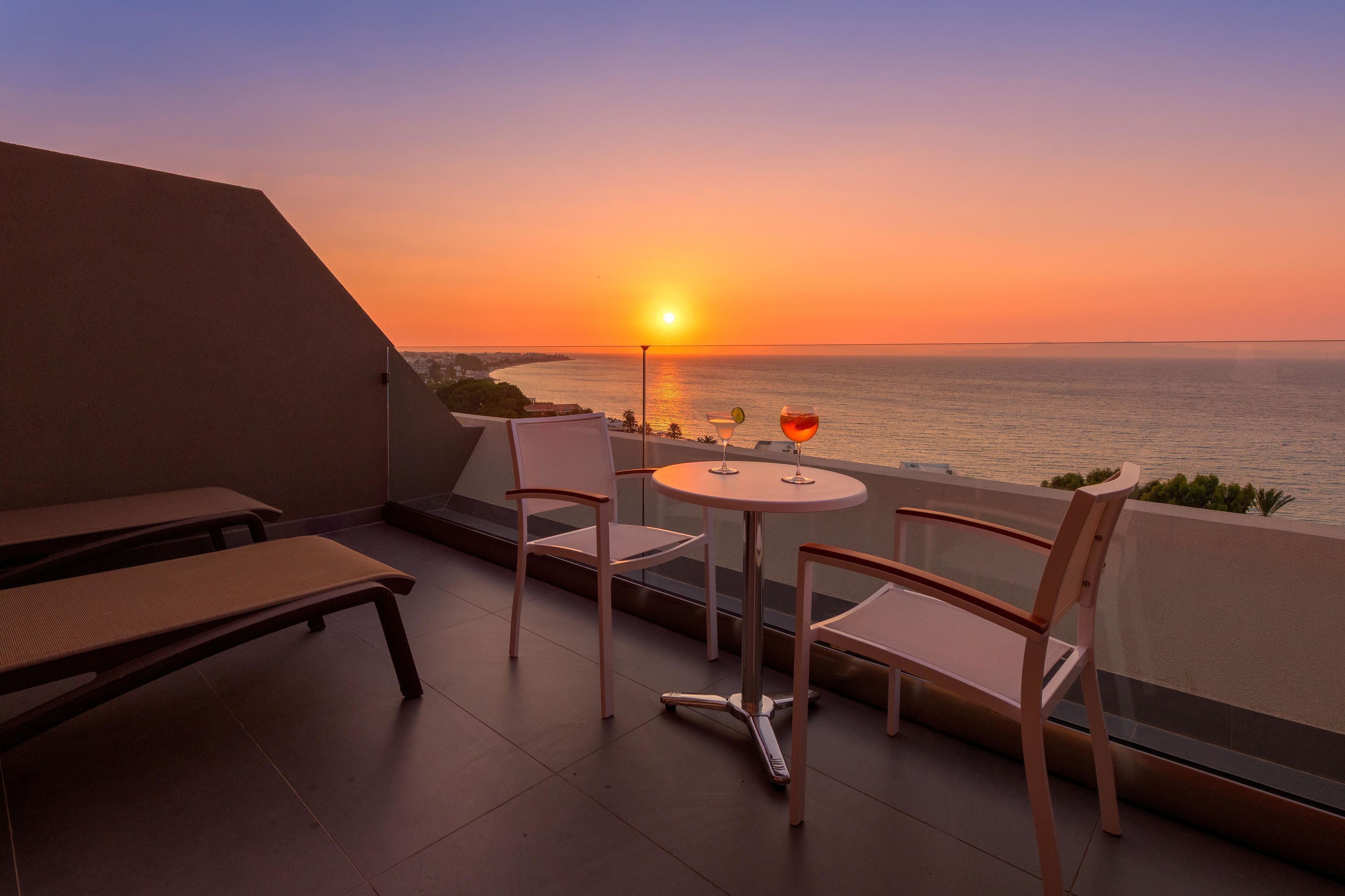 A balcony with a table and chairs, overlooking a sunset over the ocean.