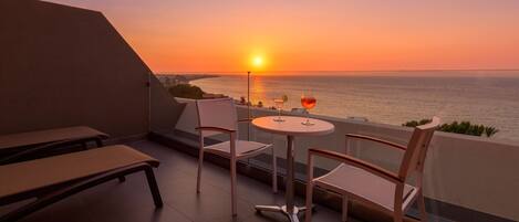 A balcony with a table and chairs, overlooking a sunset over the ocean.