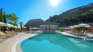 Une piscine entourée de chaises longues et d’un parasol, bordée par un bâtiment d’hôtel et avec une chaîne de montagnes en toile de fond.
